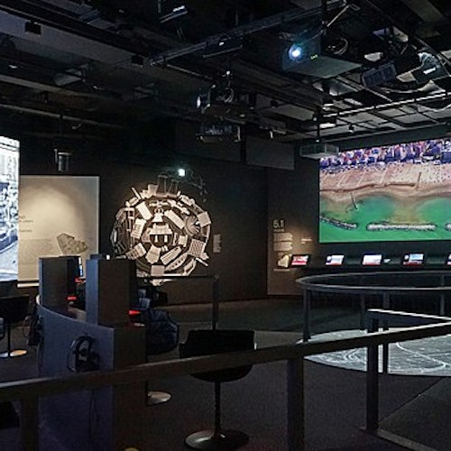 Una sala espositiva poco illuminata con display interattivi e grandi proiezioni a parete di un paesaggio urbano e di una costa.