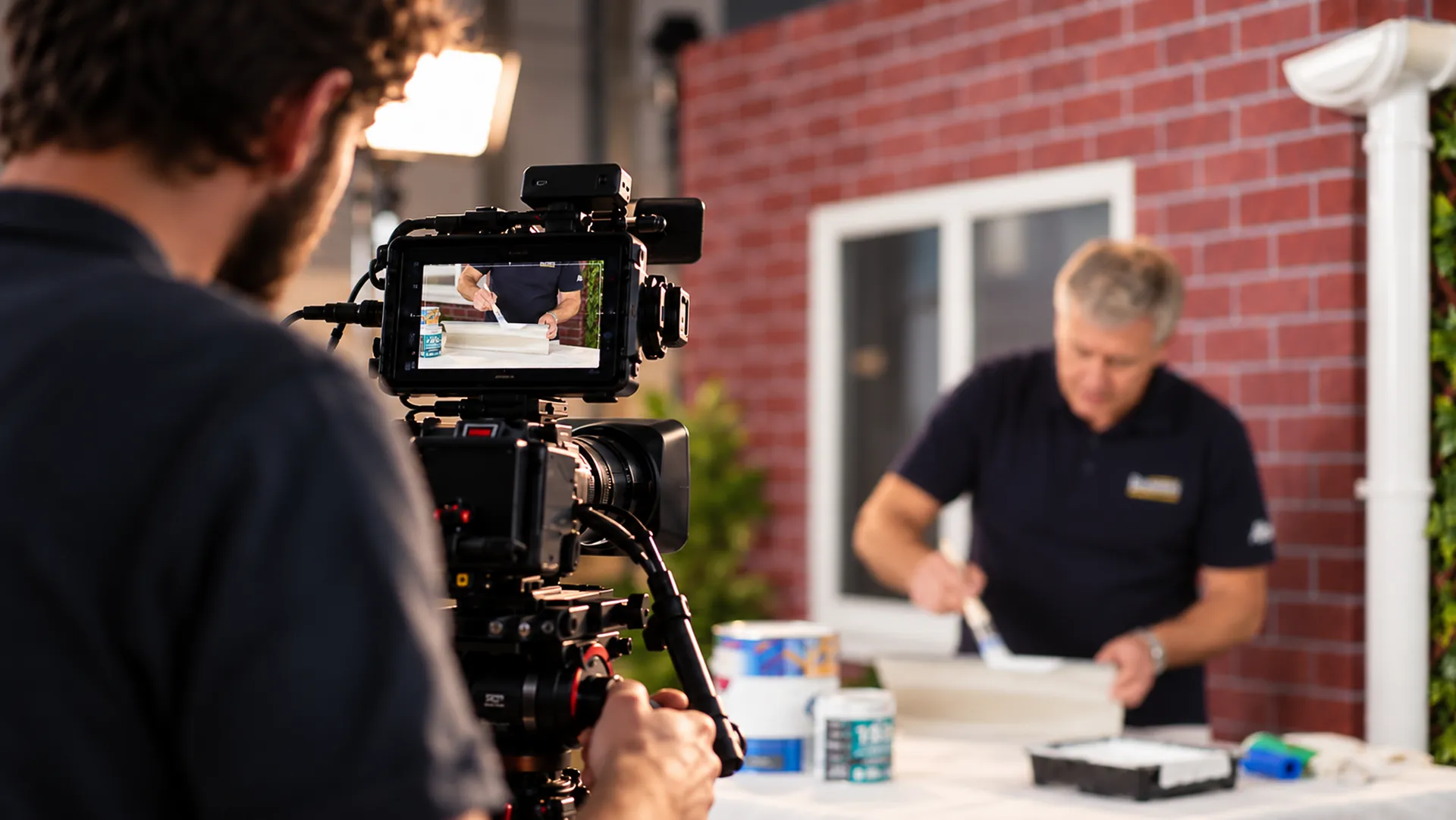  Behind-the-scenes view of a camera operator filming a demonstrator painting a section of guttering on a worktable, with shallow depth of field.