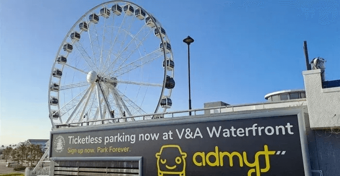 커다란 관람차를 배경으로 'Ticketless parking now at V&A Waterfront'라고 적힌 대형 옥외 광고판이 설치된 모습. 광고판 하단에는 'admyt' 로고와 자동차 아이콘이 그려져 있습니다.