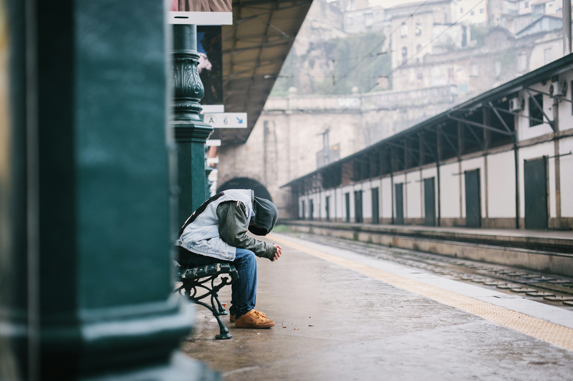 Person sitting alone on a bench at an empty, rainy train station, wearing a hooded jacket and jeans, with head down in a reflective or sad posture. The misty urban backdrop and wet platform add to the somber mood