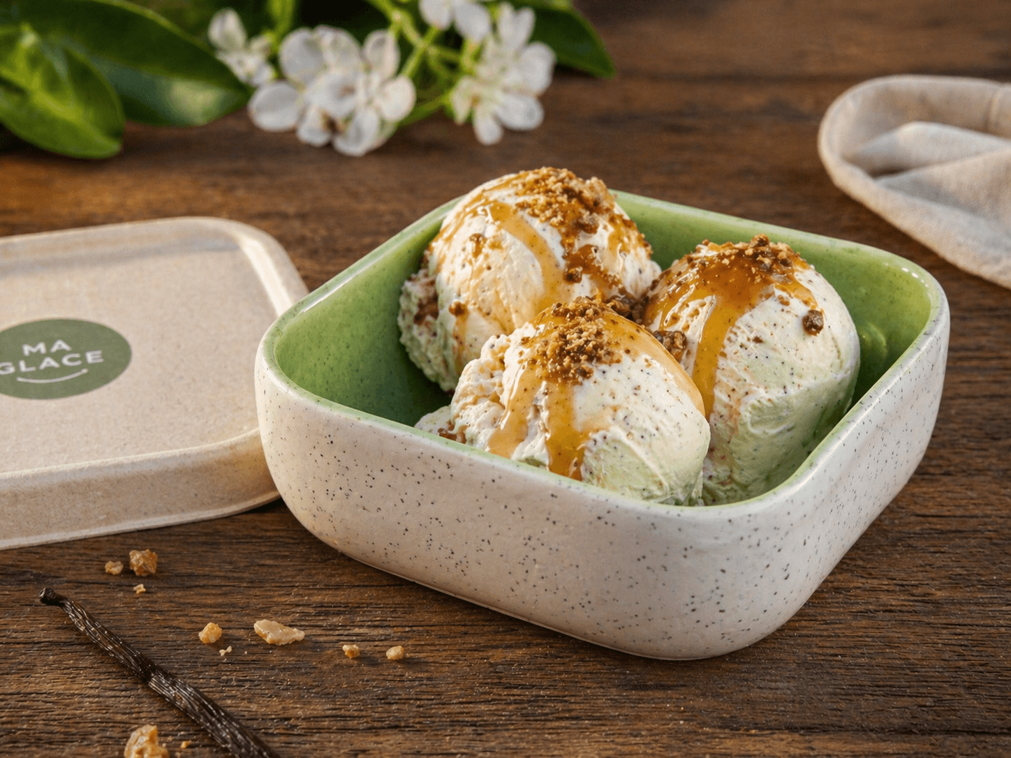 Bol carré en céramique mouchetée vert et beige garni de boules de glace vanille nappées de caramel et pralin, sur table en bois