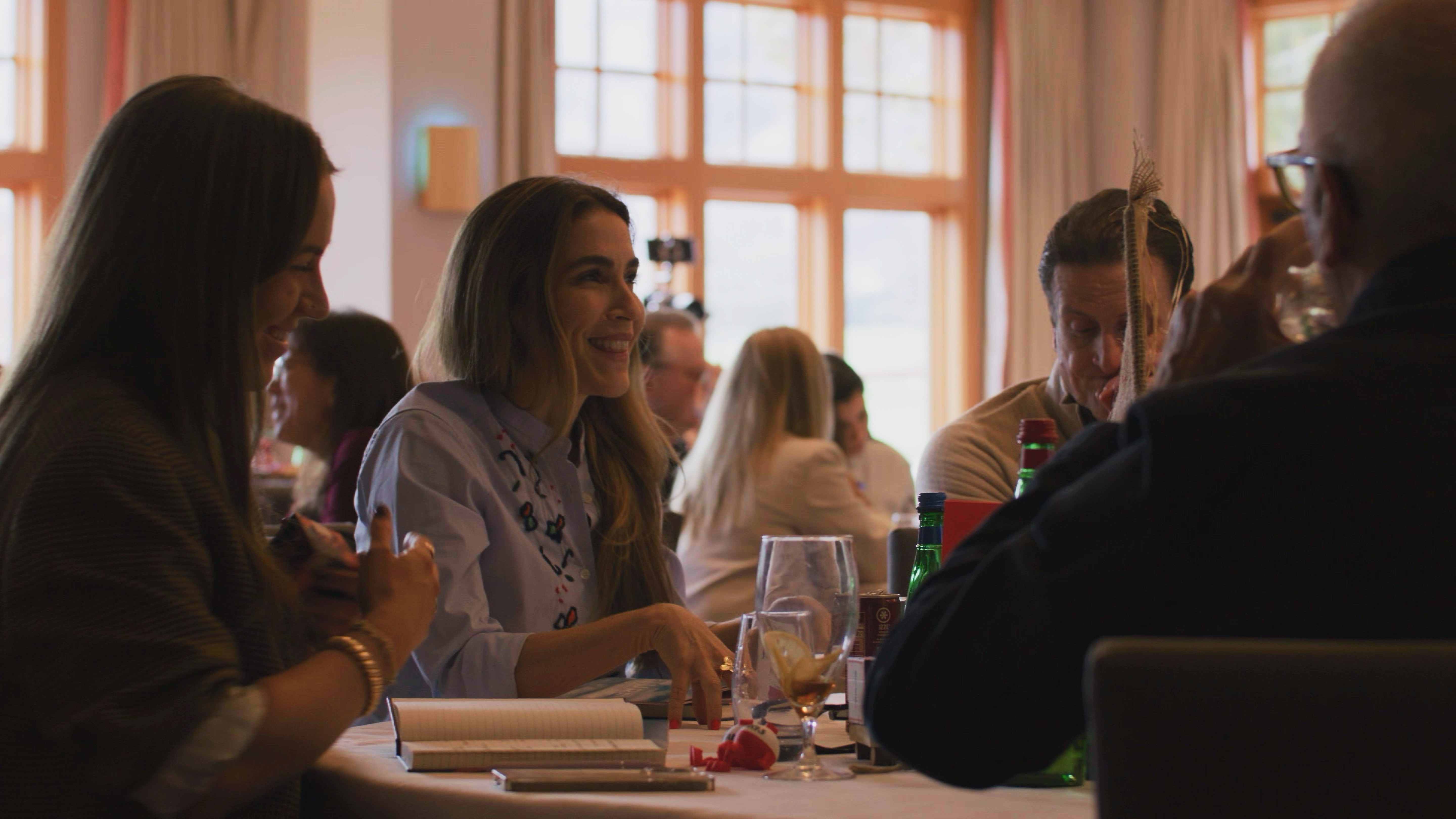 A small group of people seated around a round table in a bright indoor space, engaged in animated conversation with notebooks, papers, and cups on the table, while other groups talk at nearby tables in the background.