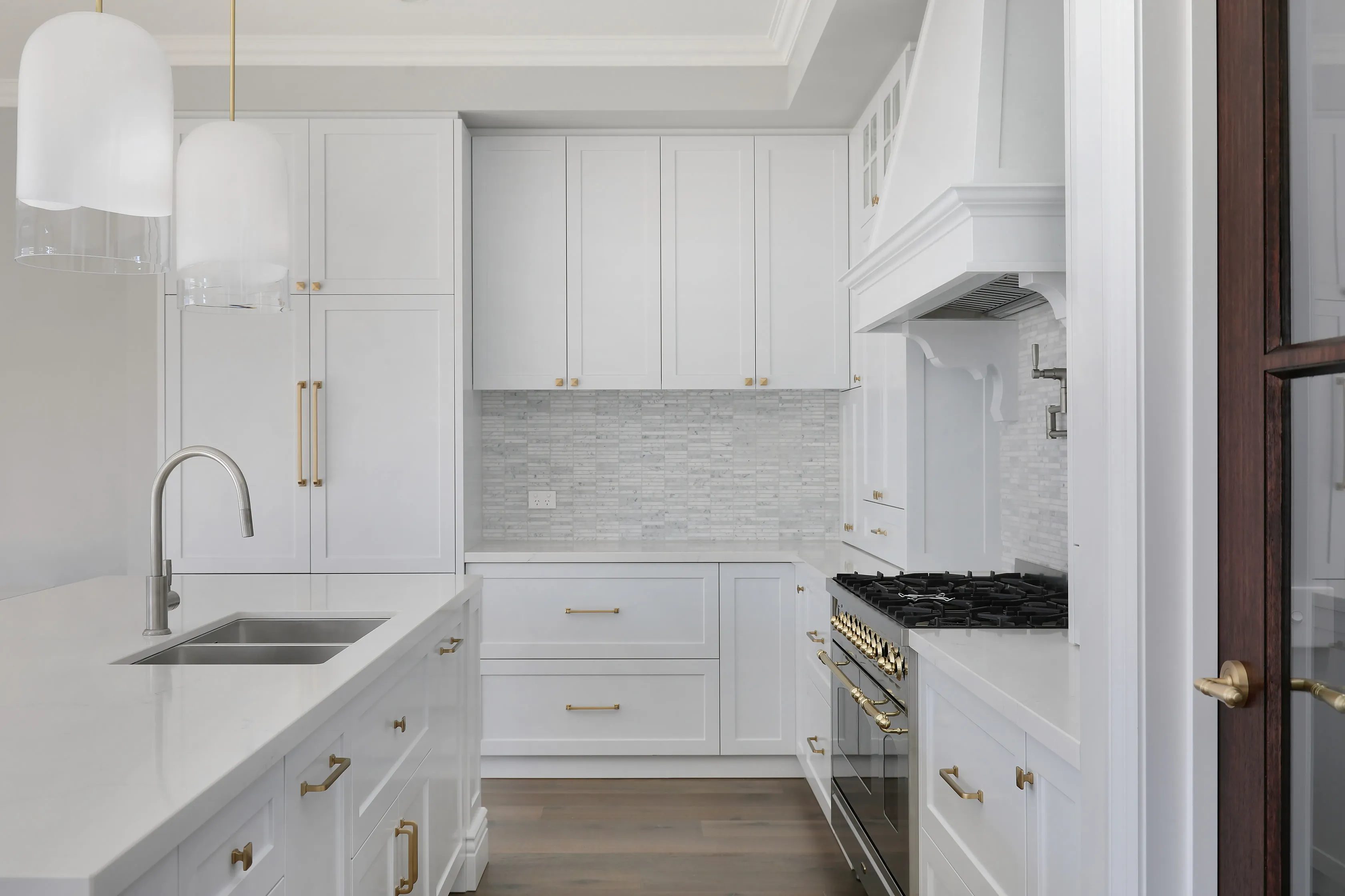 Hamptons kitchen with white shaker cabinetry and brass hardware, white dome pendants, marble mosaic splashback, a stainless and brass gas range with white mantle hood and a timber-framed glazed door