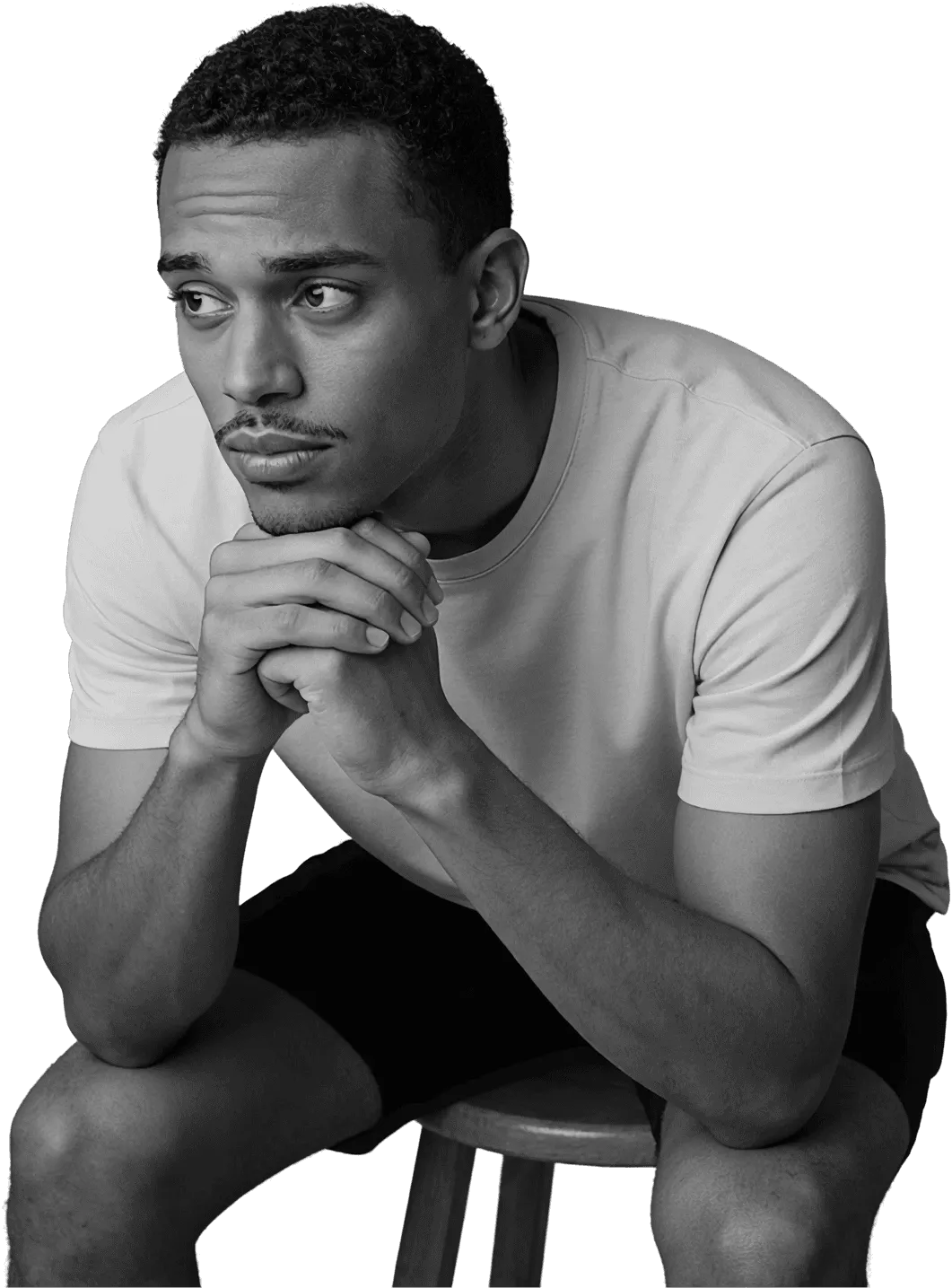 Pensive young man sitting on a stool with hands clasped under his chin, looking away in a dark studio setting