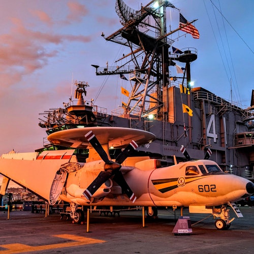 เครื่องบินทหาร หมายเลข "602" จอดอยู่บนดาดฟ้าเรือ ซึ่งมีโครงสร้างขนาดใหญ่หมายเลข "41" และธงชาติหลากหลายปรากฏอยู่ในฉากหลัง