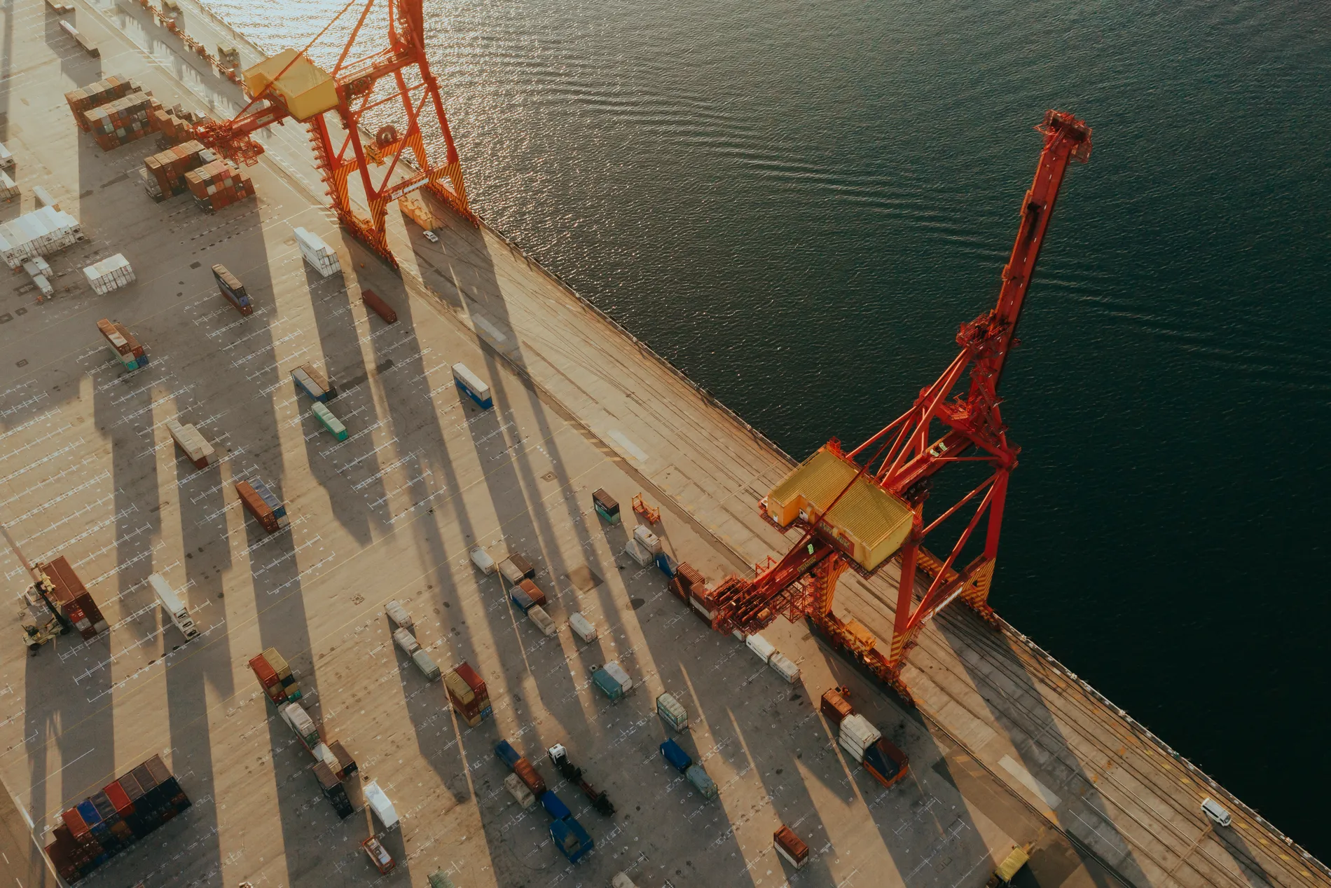 Aerial of a container terminal at golden hour, two red gantry cranes casting long shadows