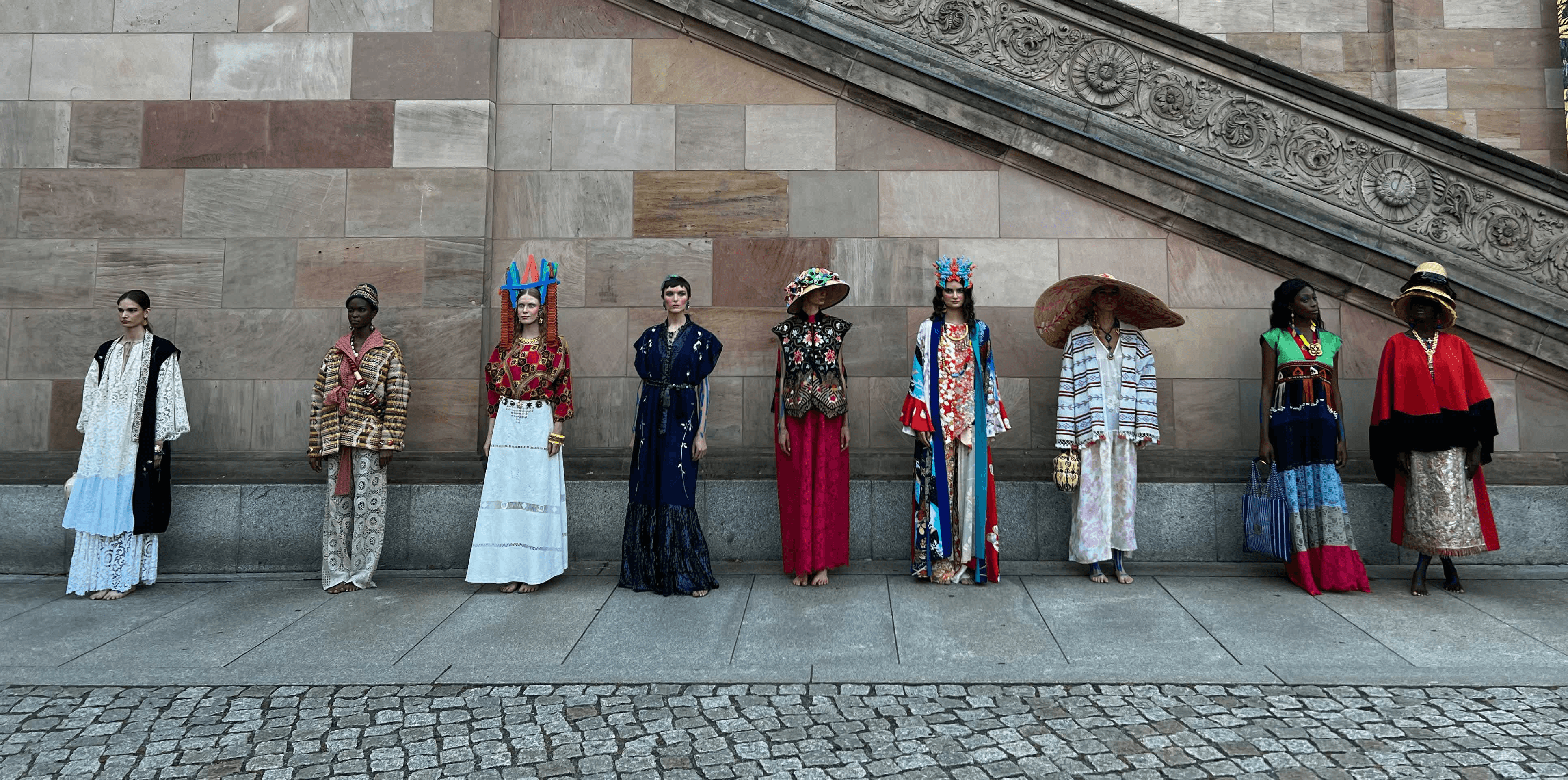 A line of nine fashion models standing side-by-side at the bottom of a wide stone staircase, showcasing various colorful and bohemian outfits from the Rianna + Nina collection.