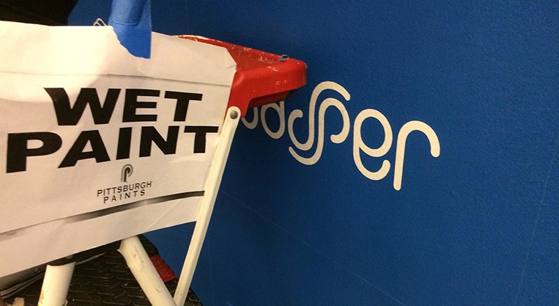 A photo with a ladder and on it a wet paint sign, near a fresh painted logo on a blue wall.
