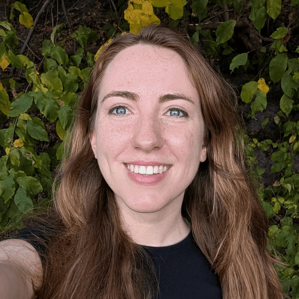 A photo of a woman with brown hair and blue eyes smiling at the camera with trees in the background