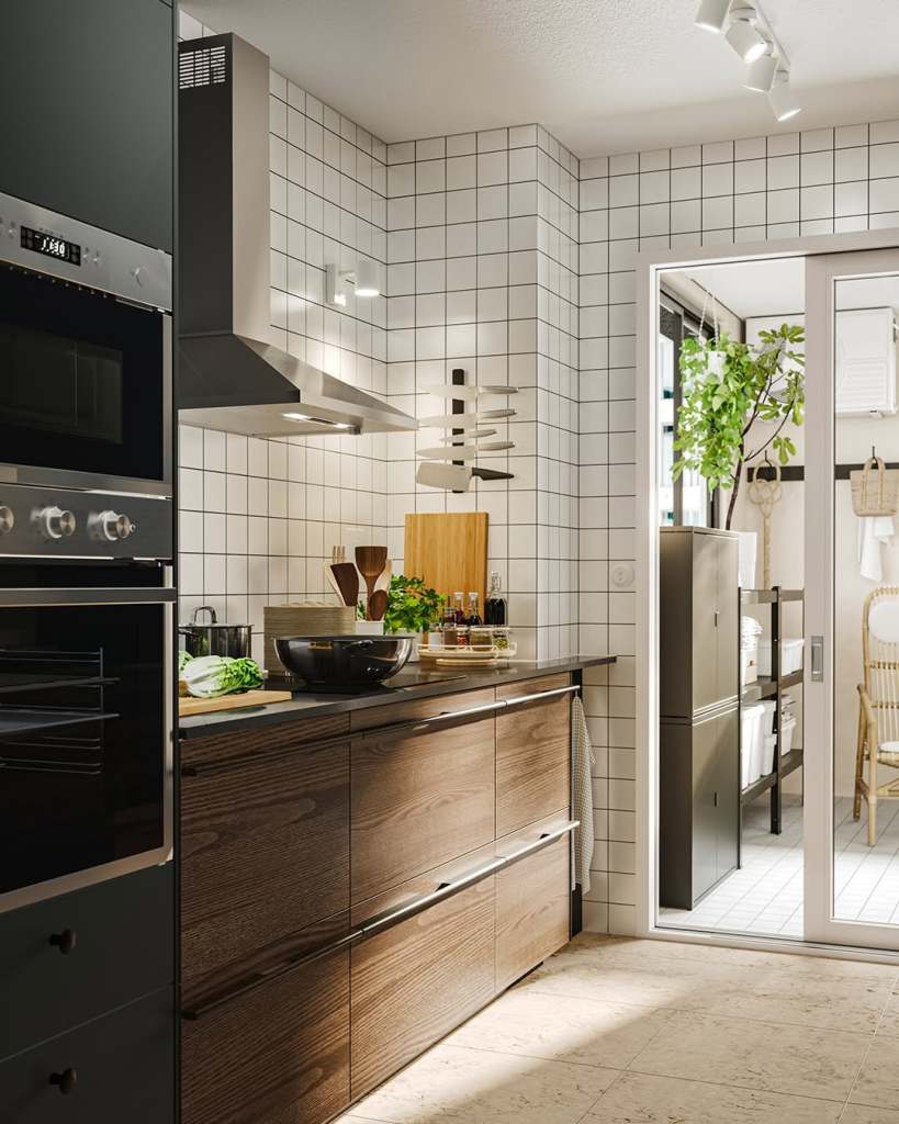 A modern kitchen featuring dark wooden cabinets, a black countertop, stainless steel appliances, and a tiled backsplash. The kitchen is bright and airy, with a doorway leading to a well-organized pantry area filled with plants and kitchen items.