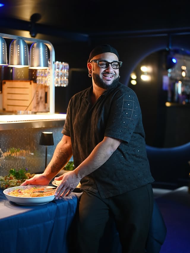 A smiling man with a beard and glasses, wearing a black patterned short-sleeve shirt and cap, is serving himself food from a buffet line under warming lamps.