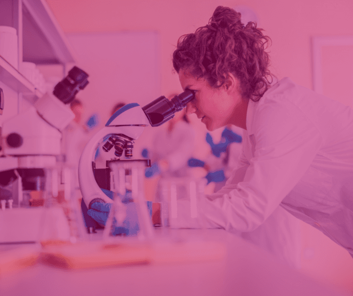 Uma cientista está em um laboratório, observando através de um microscópio. Ela usa luvas azuis e um jaleco branco, em um ambiente repleto de equipamentos científicos e frascos de vidro sobre a bancada. O fundo está desfocado, mostrando outras pessoas no laboratório, sugerindo um ambiente de pesquisa colaborativo. A imagem tem uma tonalidade rosada, destacando a presença feminina na ciência.