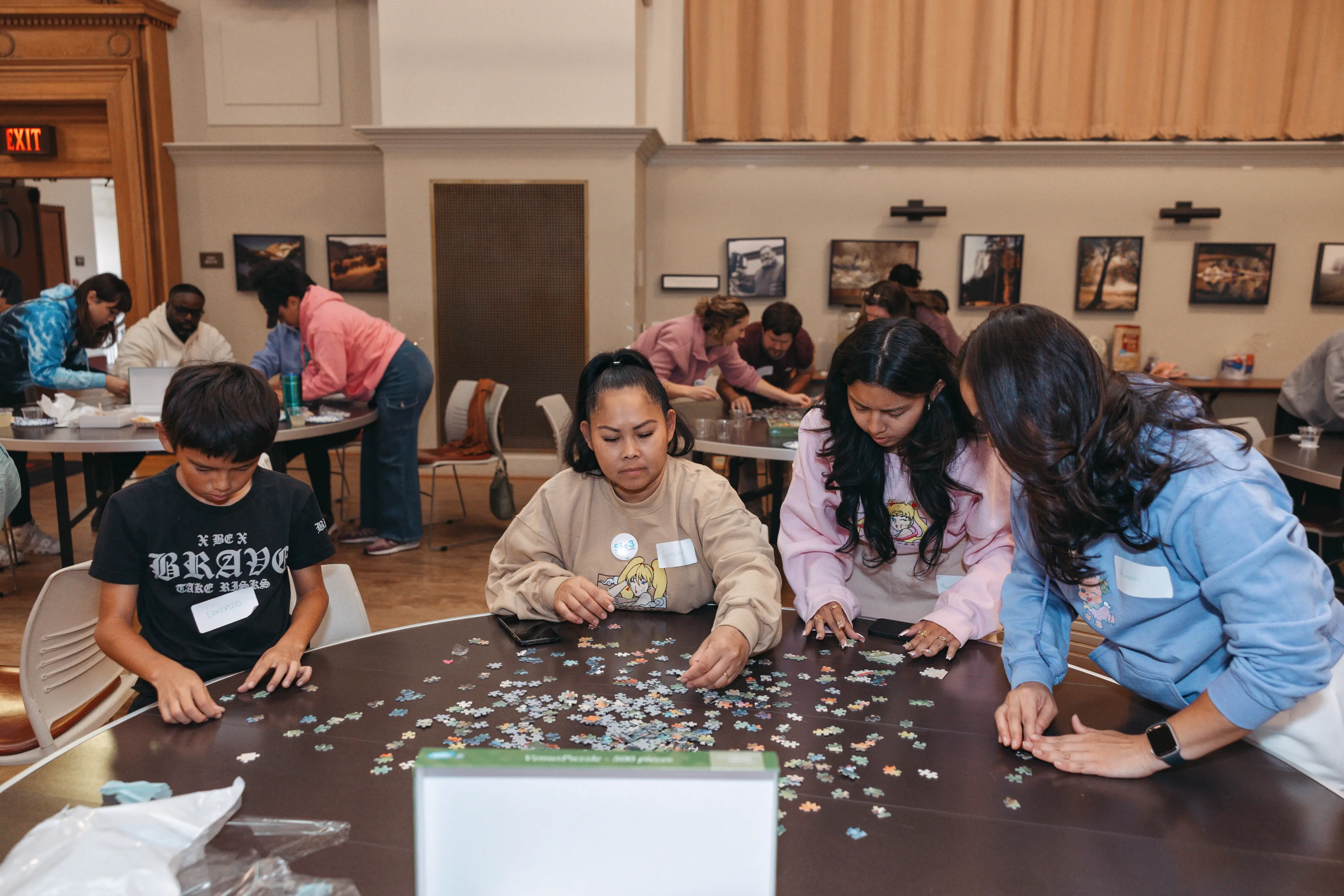 Un grupo de personas, incluidos niños y adultos, está concentrado en armar un rompecabezas en una mesa redonda en una habitación bien iluminada con arte en las paredes, lo que sugiere un evento comunitario o de integración de equipo.