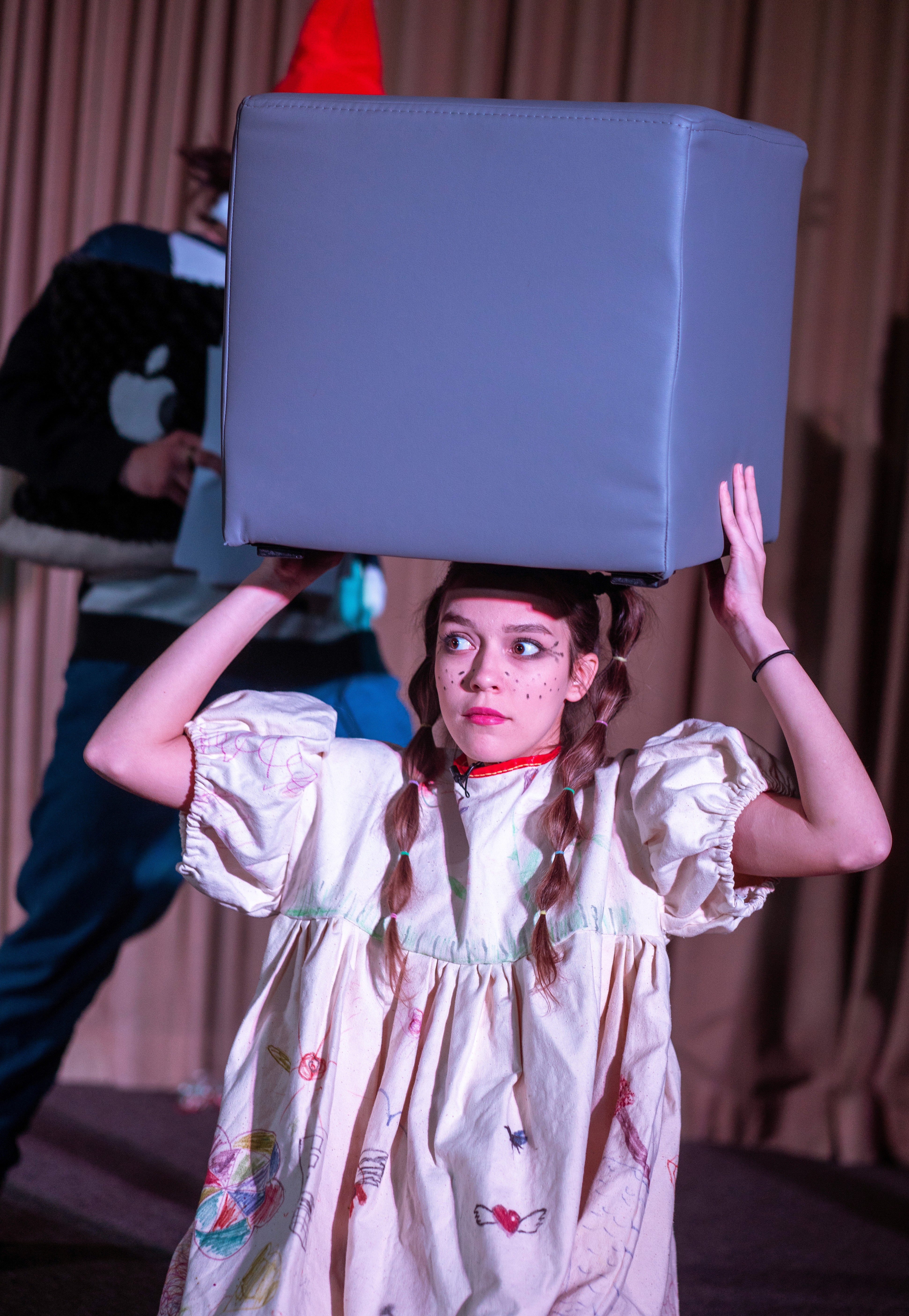 Foto der jungen Frau, die in dem Theaterstück auftritt, in einem weißen Kleid und mit einem großen grauen Würfel auf dem Kopf.
