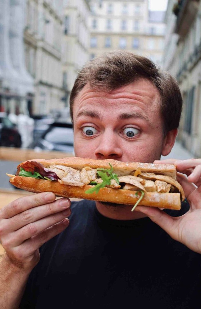 Une personne tient un grand sandwich à la baguette garni de poulet grillé, de légumes croquants et de légumes frais, tout en se tenant dans une rue animée de la ville, avec des bâtiments historiques en arrière-plan.