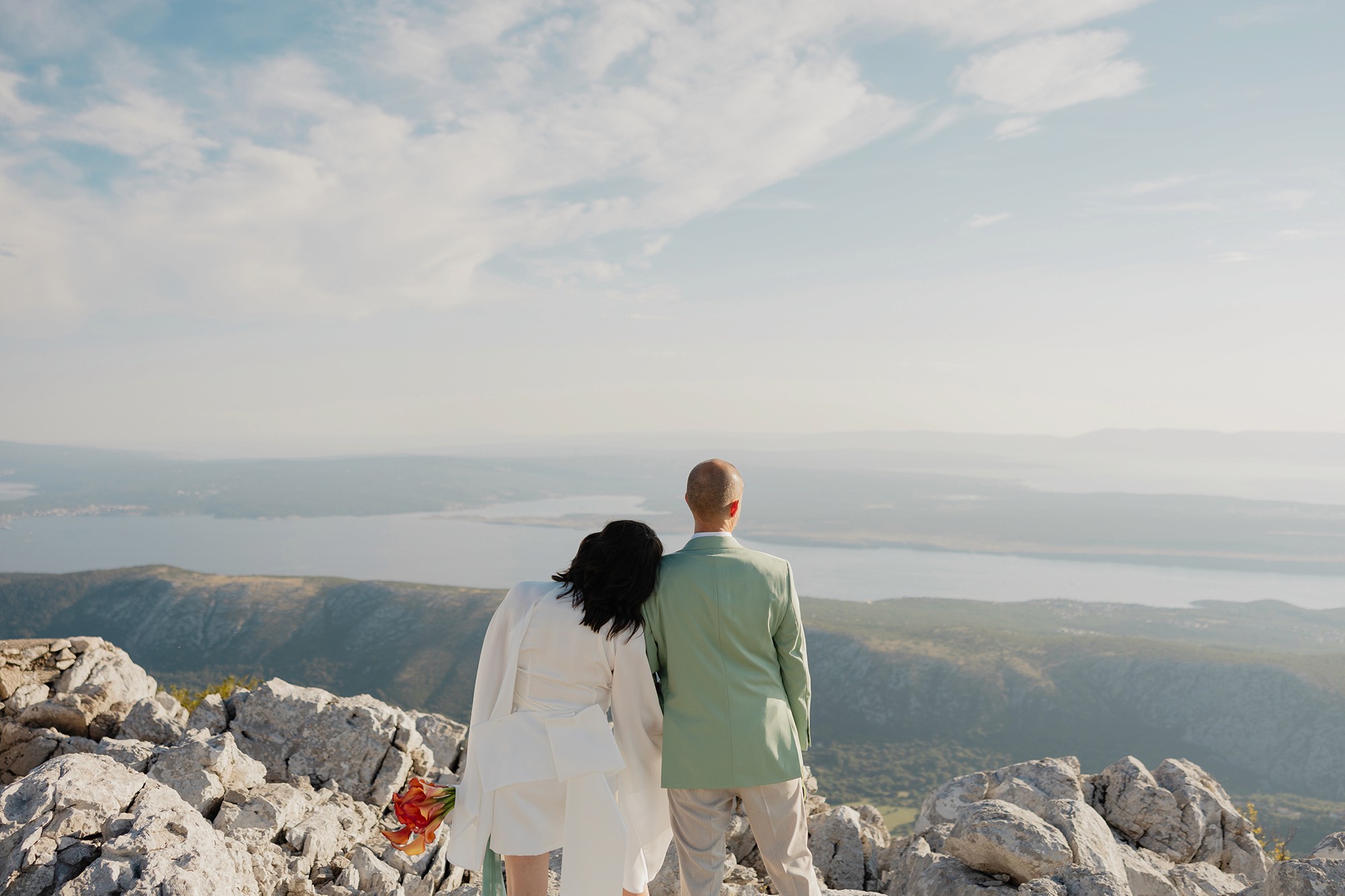 Newly wed couple standing whilst staring out at a clifftop view of sea and island