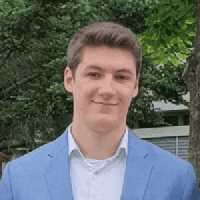 A young man wearing a blue suit jacket over a white shirt, standing confidently with a neutral expression.