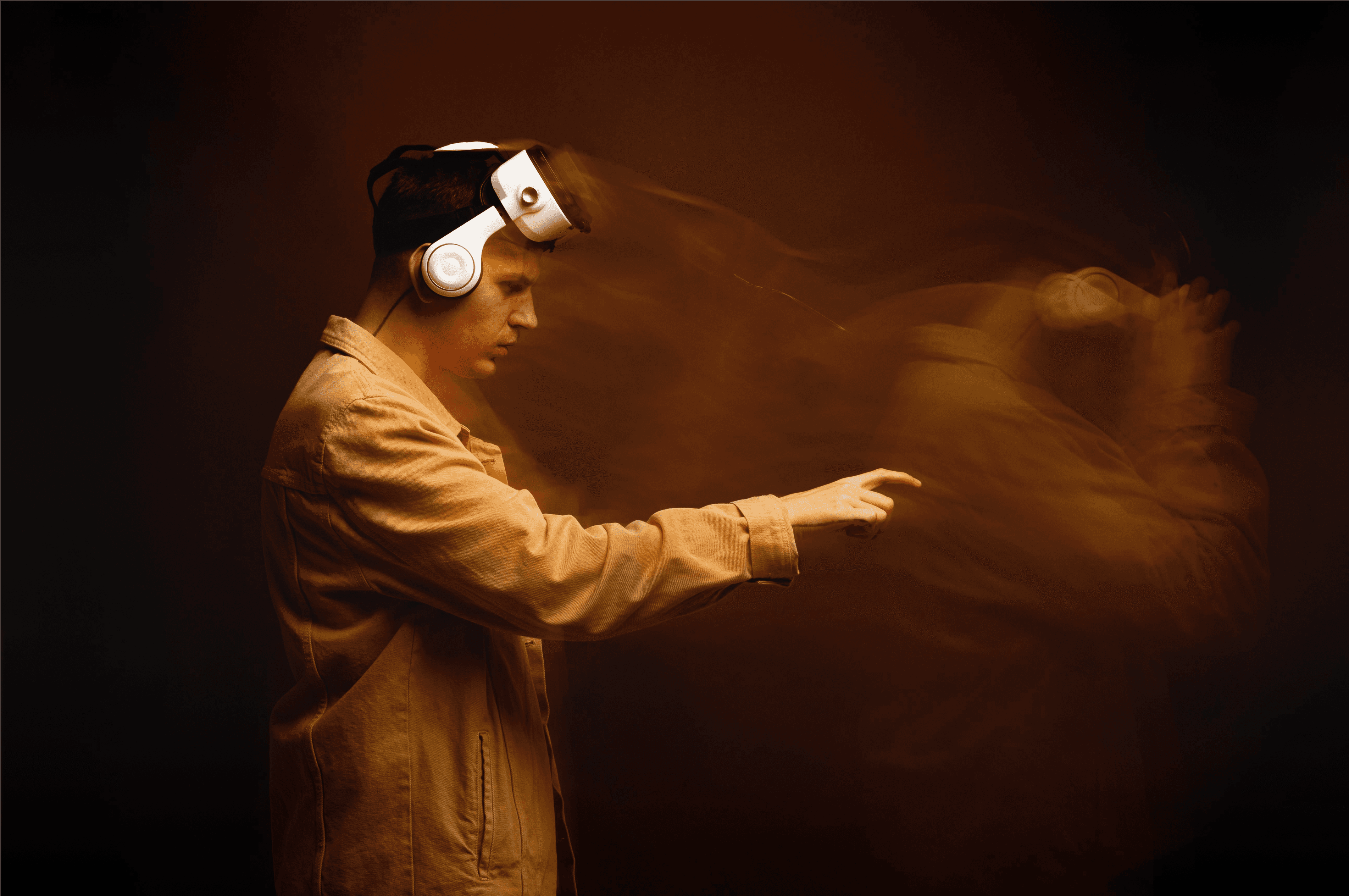A cinematic side-view portrait of a man wearing a white VR headset and a mustard yellow jacket. The image features a motion blur effect, showing a trailing ghostly silhouette of the person moving forward, set against a dark amber background with warm, moody studio lighting.