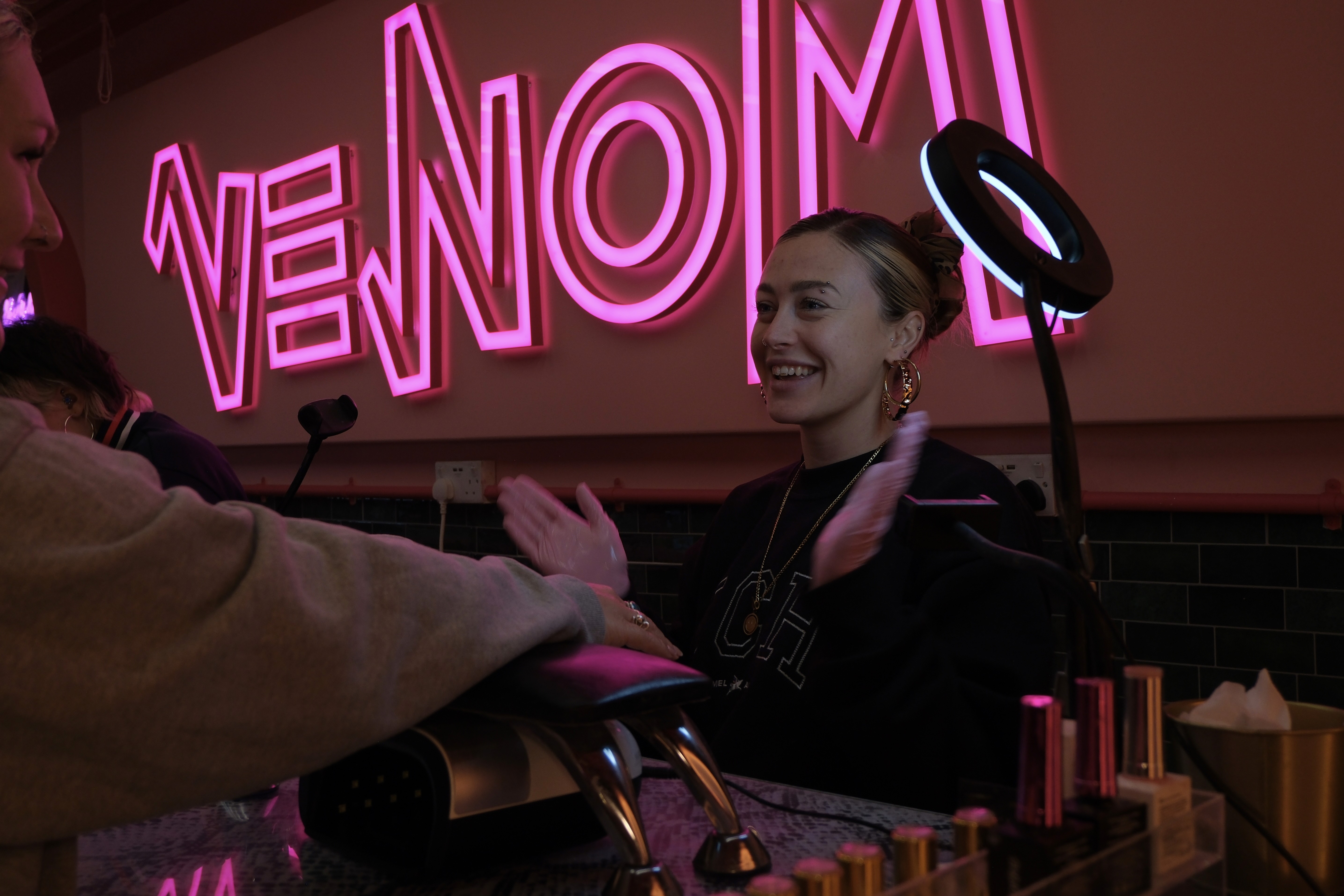 A wide, candid shot of a smiling nail technician wearing gold hoop earrings and pink nitrile gloves, mid-conversation with a client. Her hands are slightly blurred in motion, adding a dynamic feel to the scene. The glowing pink neon "VENOM" sign serves as a bold backdrop, with professional tools, a ring light, and various gel polish bottles visible on the snakeskin-patterned desk in the foreground.