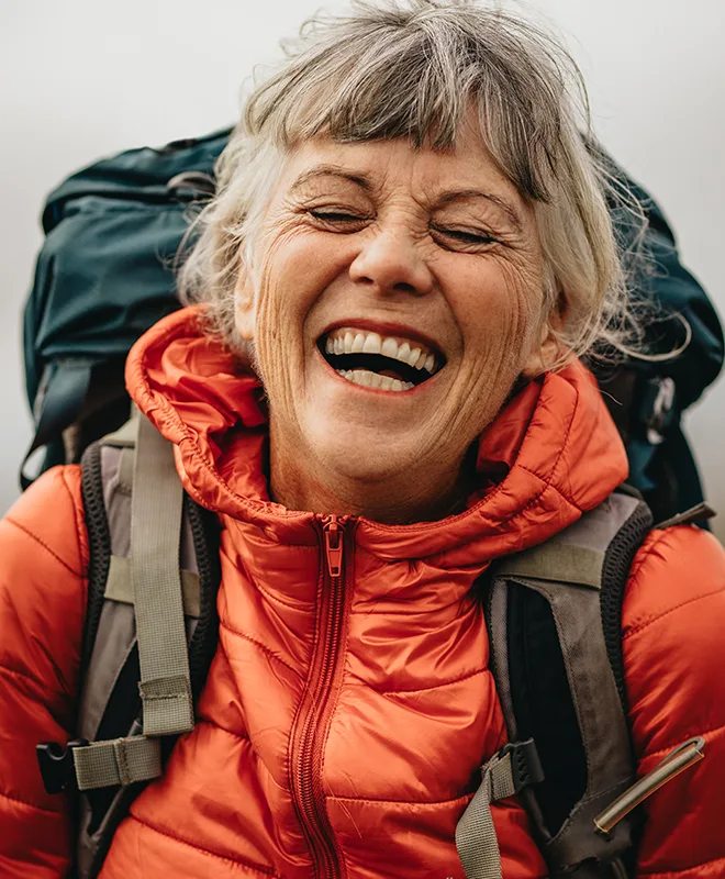 An older woman in a red puffer jacket laughing joyfully while hiking with a backpack