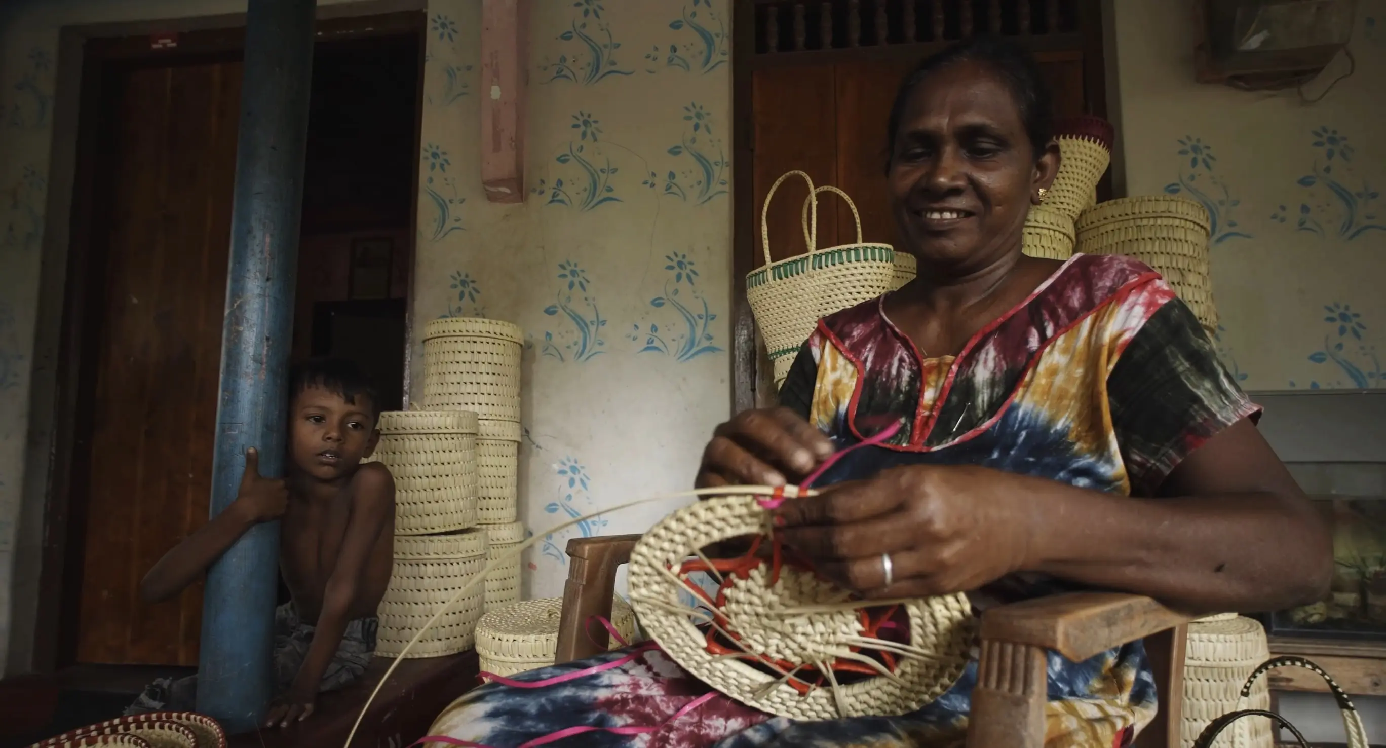A woman working on weaving a basket with a child pondering on the side