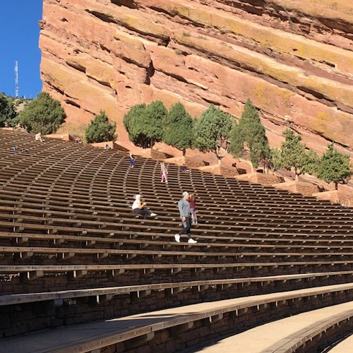 People walking and sitting on tiered stone seating with a backdrop of large red rock formations and sparse trees under a clear blue sky.