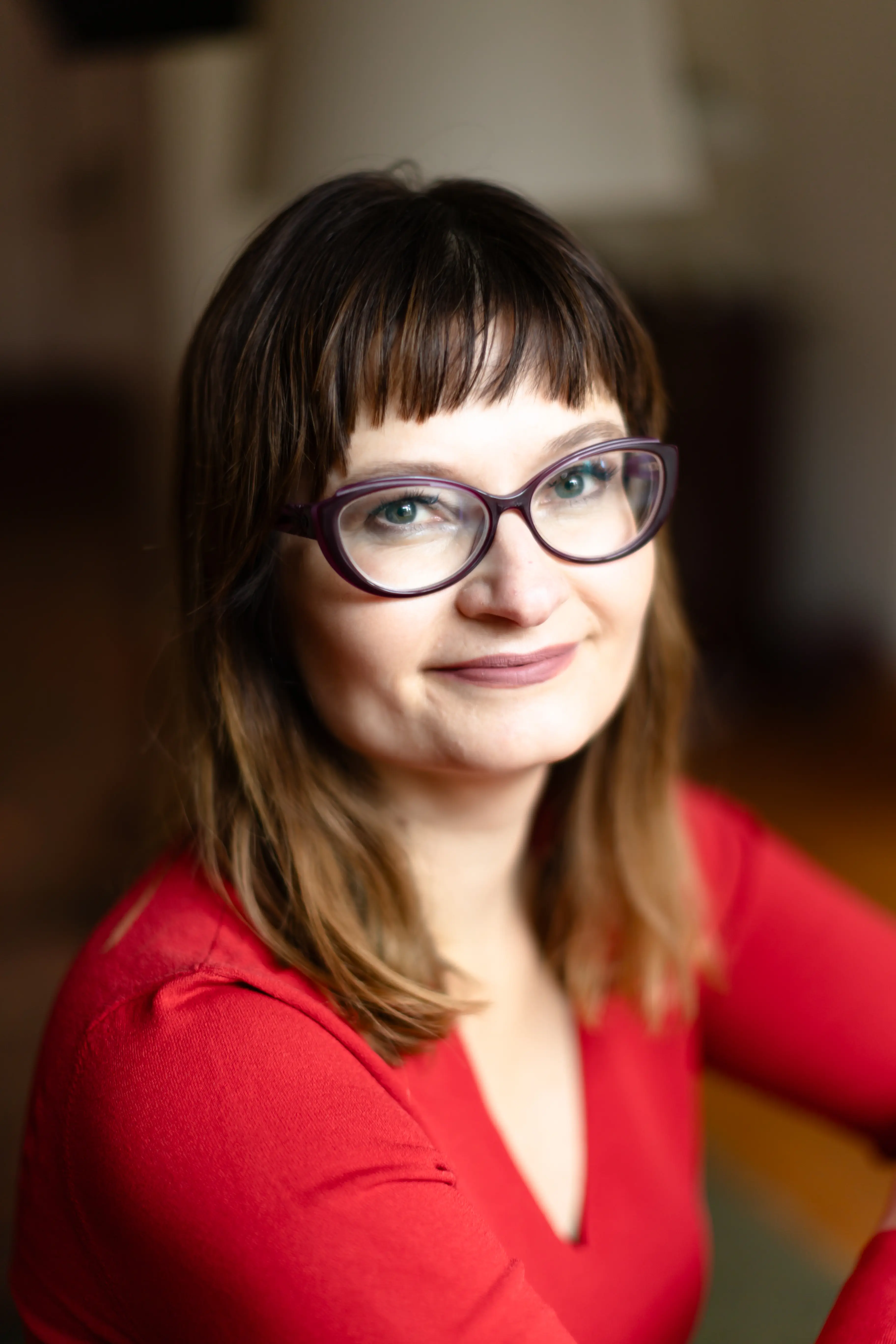 Kobieta z blond-brązowymi włosami i okrągłymi okularami, uśmiecha się w jasnej czerwonej bluzce. Tło jest rozmyte, z dużym lampą w tle.