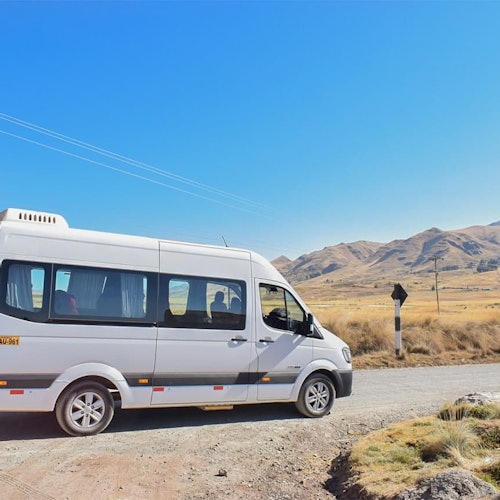 Ένα λευκό μικρό λεωφορείο σε έναν χωματόδρομο σε αγροτική περιοχή με βουνά και έναν καθαρό μπλε ουρανό στο βάθος.