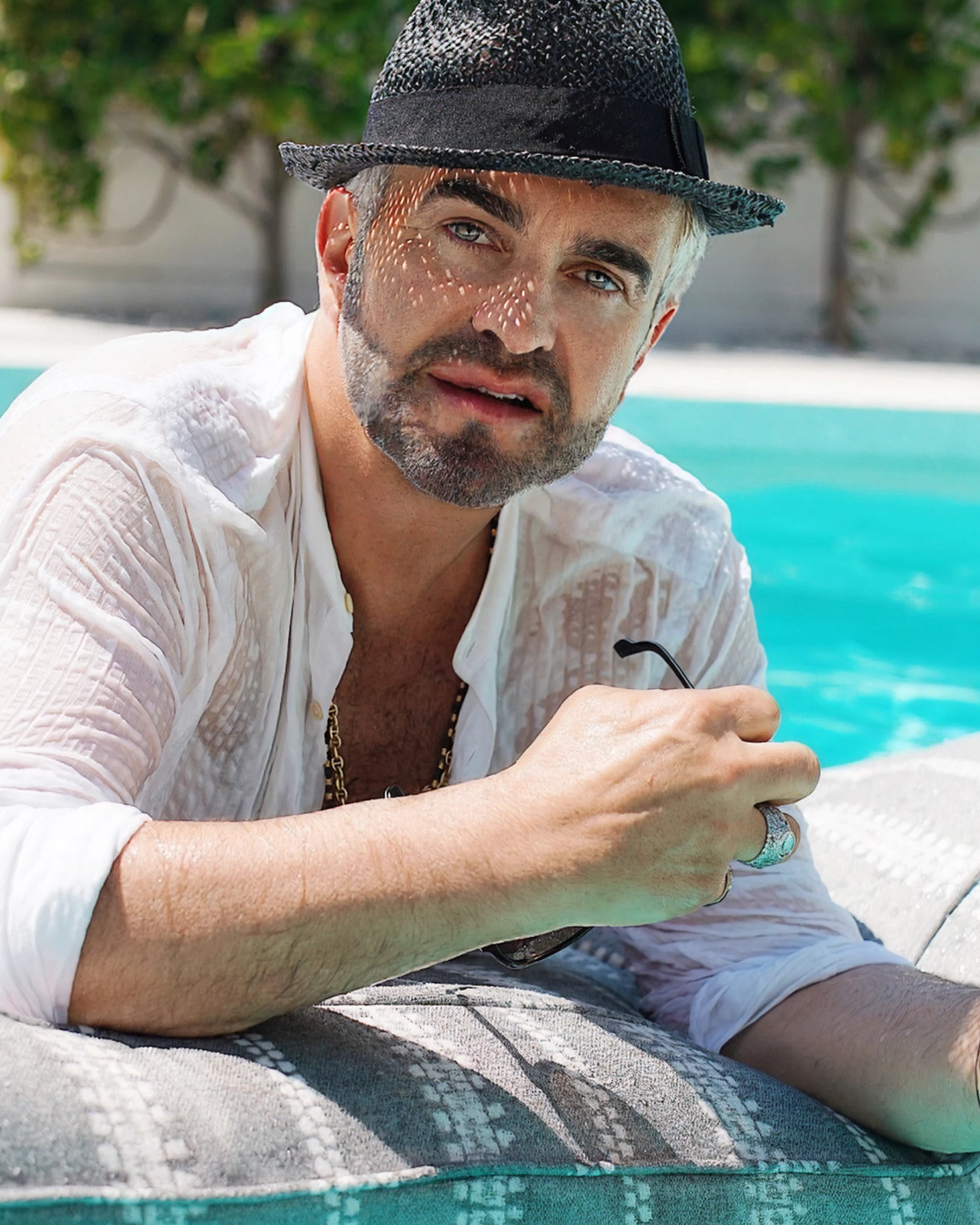 Stylish man in a black hat and white shirt relaxing poolside with Oliver James Lilos — capturing the spirit of slow luxury and effortless confidence.