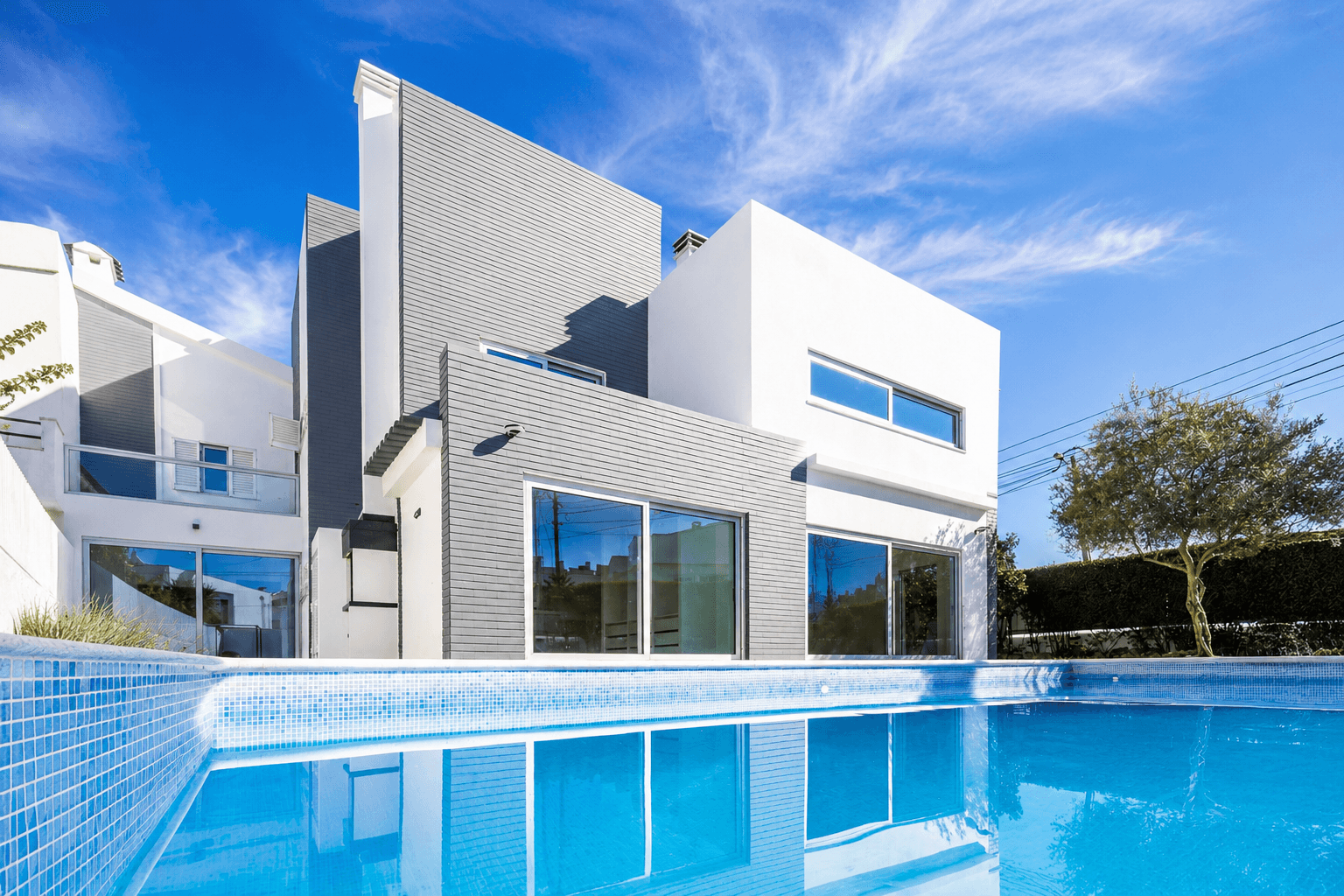 A luxurious two-story white villa with smooth, rounded architectural lines and wide arched openings. A large outdoor patio with seating is shaded by the building's overhang. In the foreground, a calm swimming pool reflects the warm tones of the architecture and the soft daylight, surrounded by minimalist landscaping and olive trees.