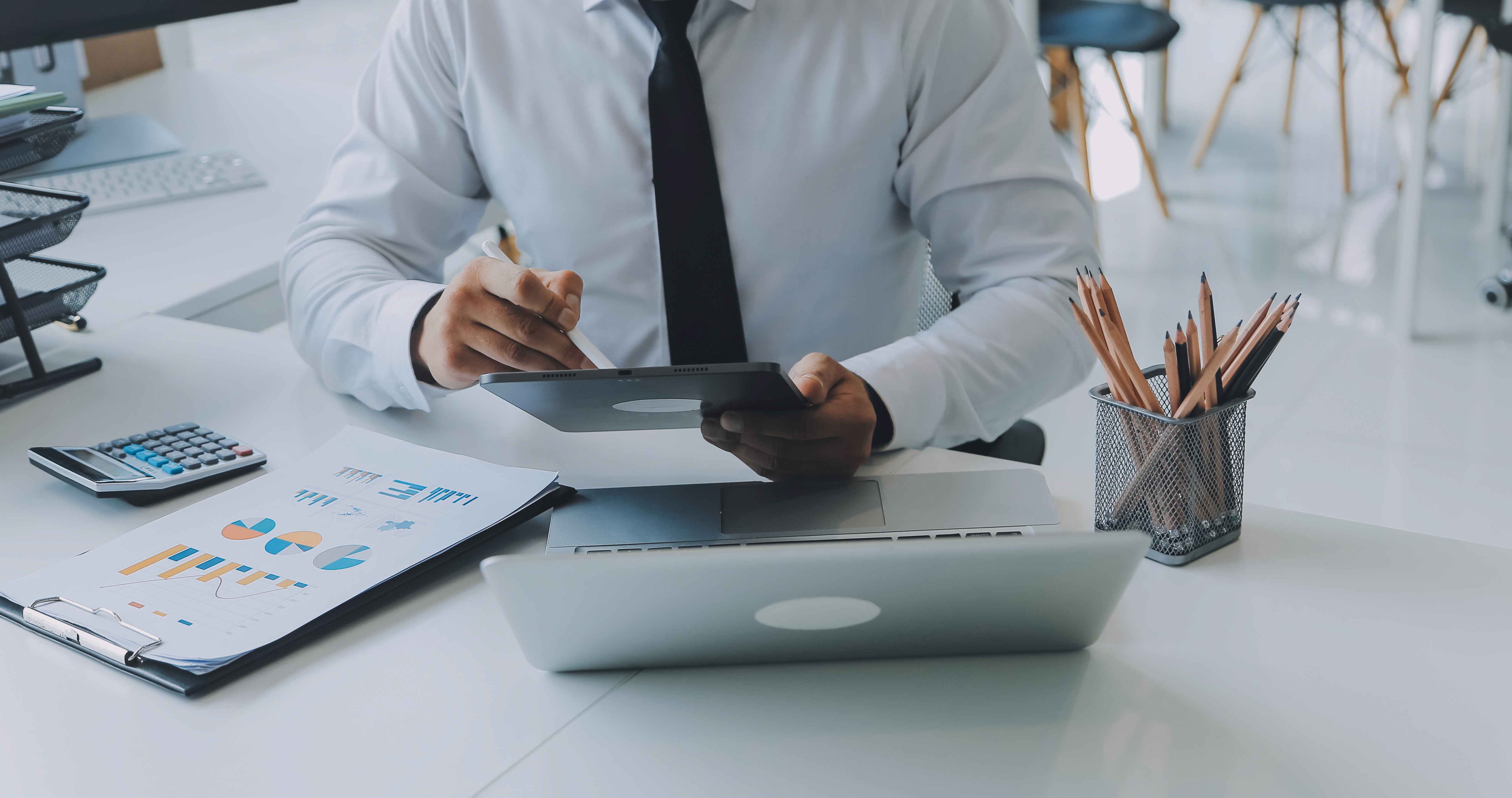 A man wearing glasses and a blazer working at a desk, writing in a notebook beside a computer monitor in a modern office.