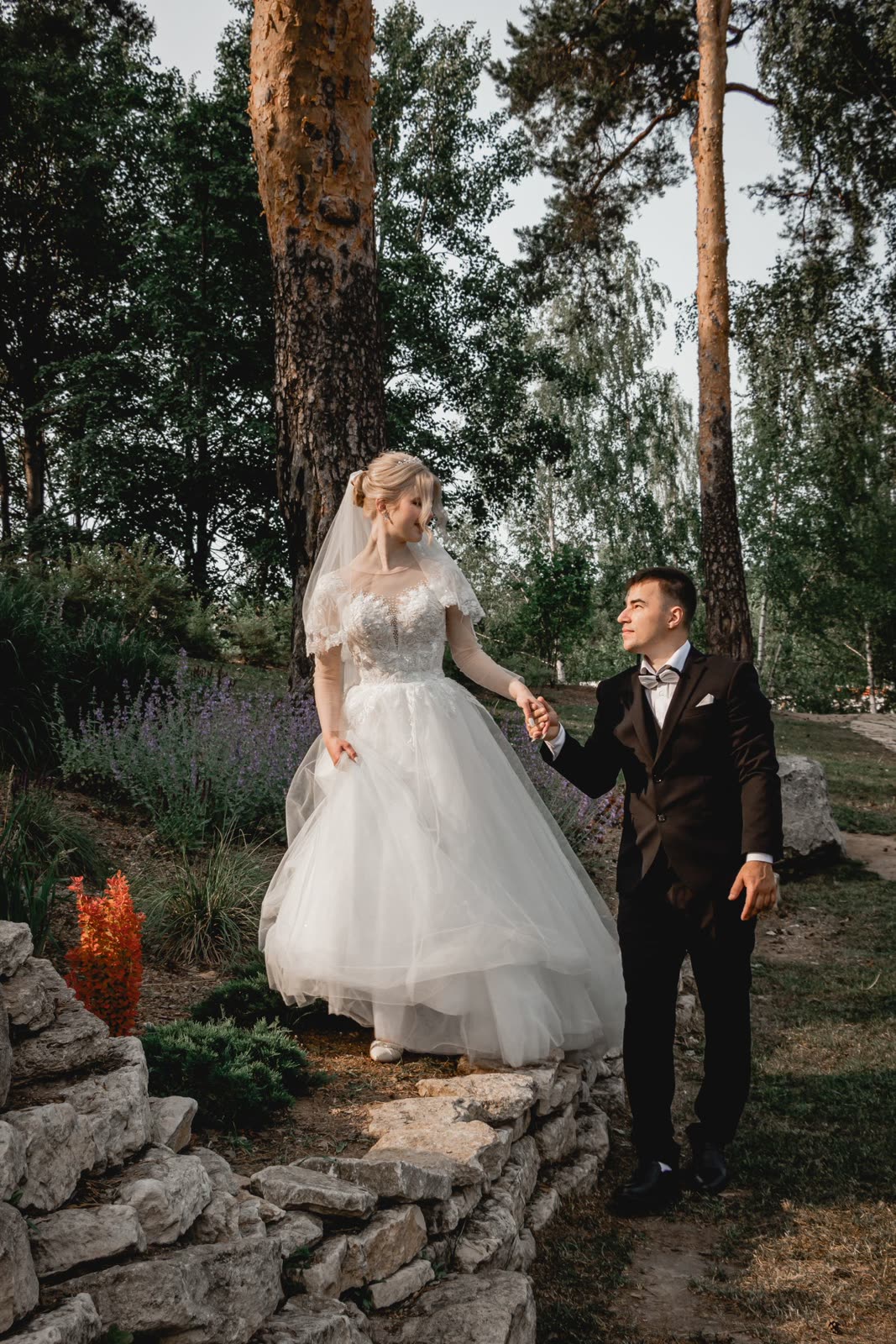 Bride in a long gown holding the groom's hand and walking on a garden path