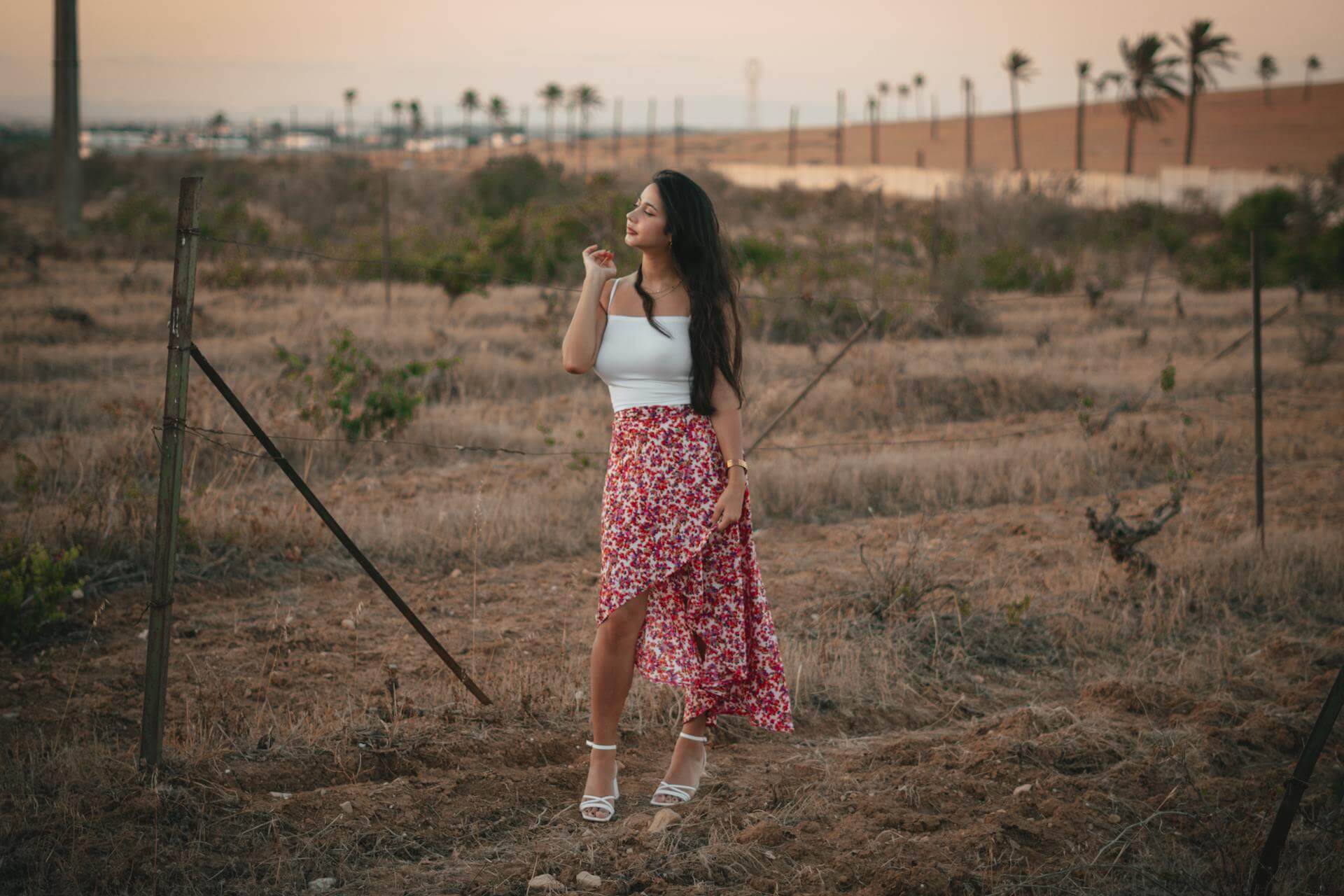 A woman in a white top and floral skirt stands gracefully in a vibrant green field under a clear blue sky