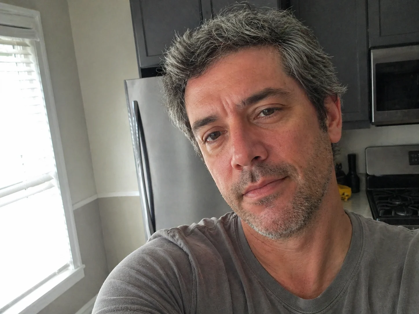 Casual kitchen selfie of a man with salt-and-pepper hair in soft natural daylight.