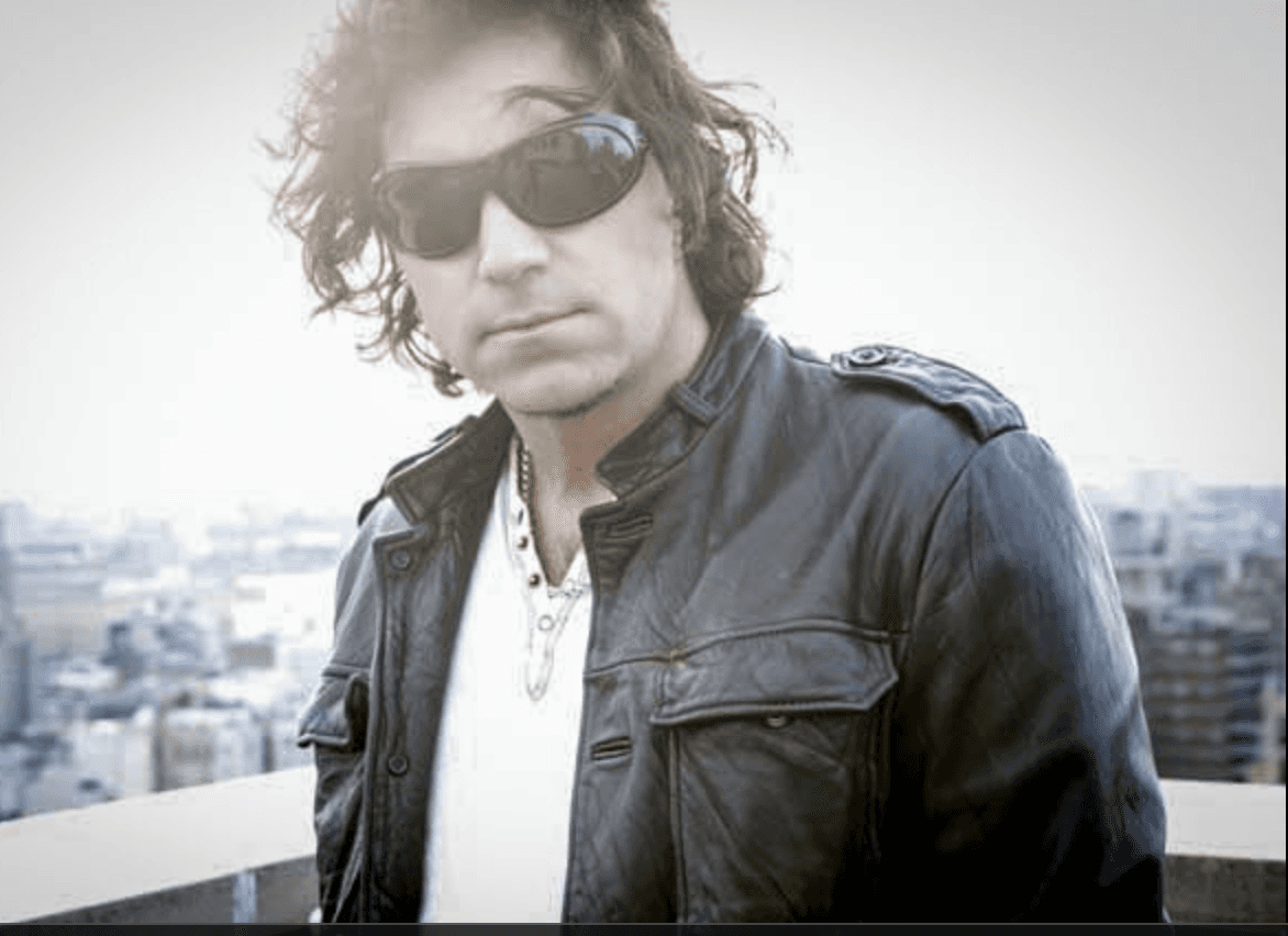 Director Kennedy, known for his work in sports, fashion, and music videos, stands on a rooftop overlooking the city. He wears a black leather jacket, dark sunglasses, and a white shirt with layered necklaces, exuding a confident, rock-influenced style.