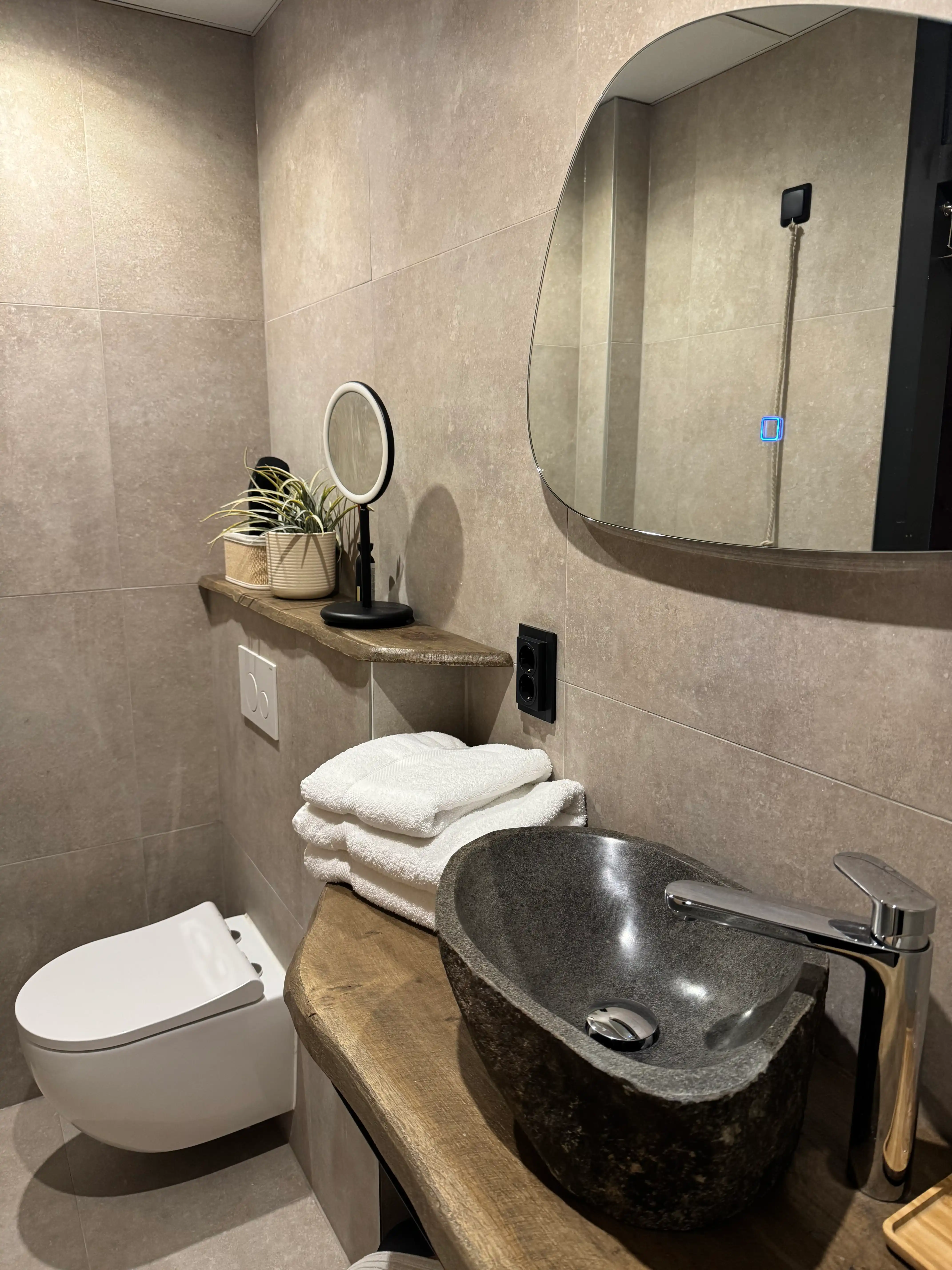 Modern zwevend toilet met beige tegels en houten wandplank.