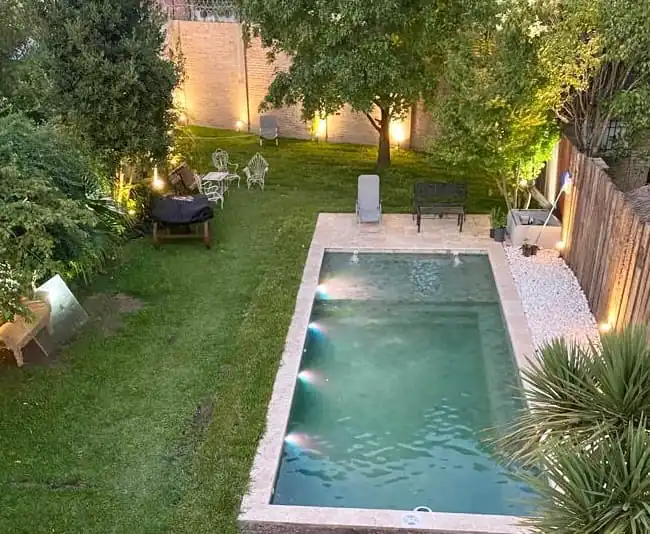 ista aérea de una piscina de hormigón iluminada de noche con luces LED perimetrales, rodeada de un amplio jardín con árboles y un sendero de piedras blancas.