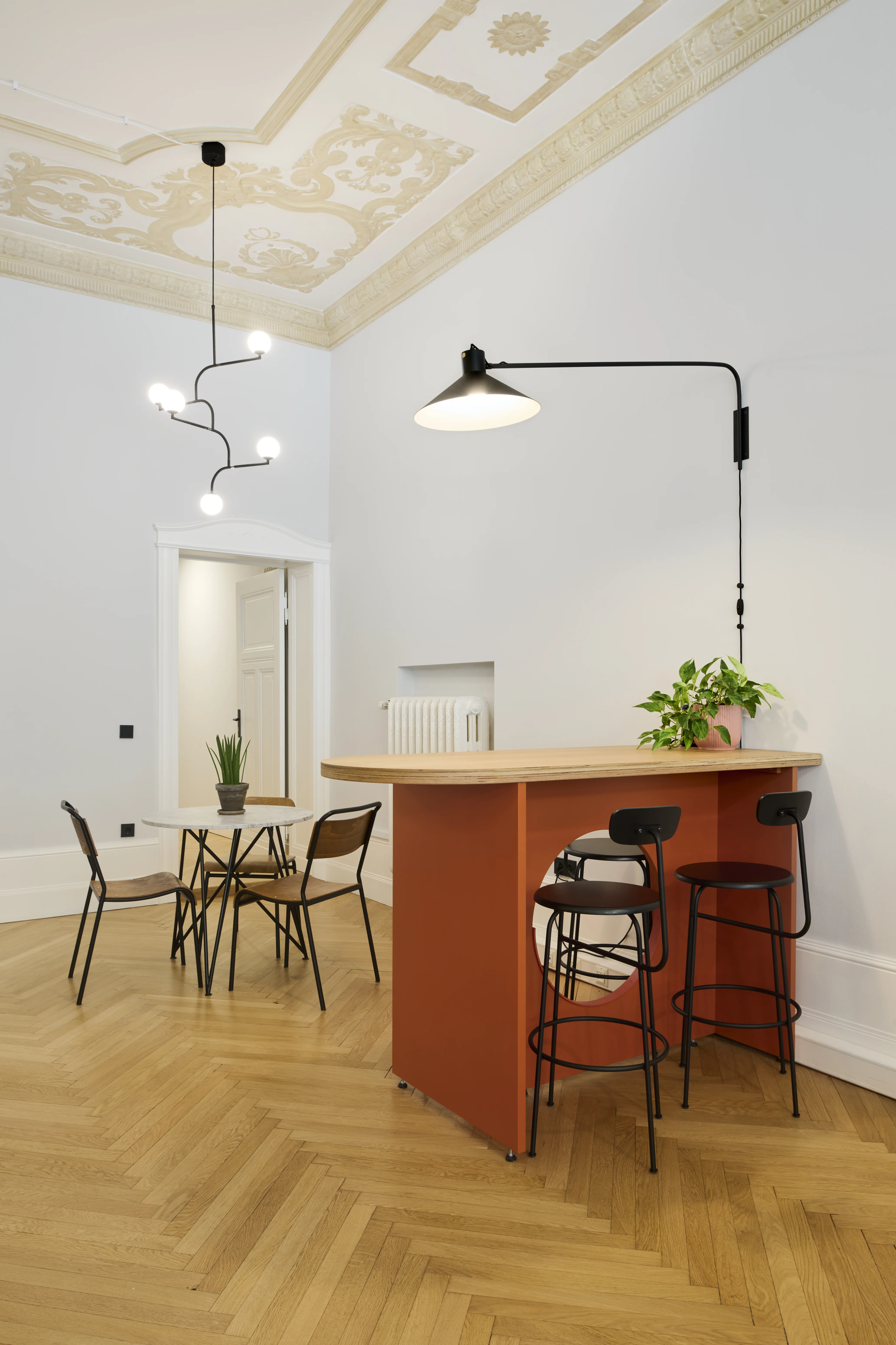 Kitchen corner at INTERMATE BERLIN with a terracotta-red bar counter with oak top, black bar stools, a black swing-arm wall lamp, a mobile light sculpture and gilded stucco ceiling.