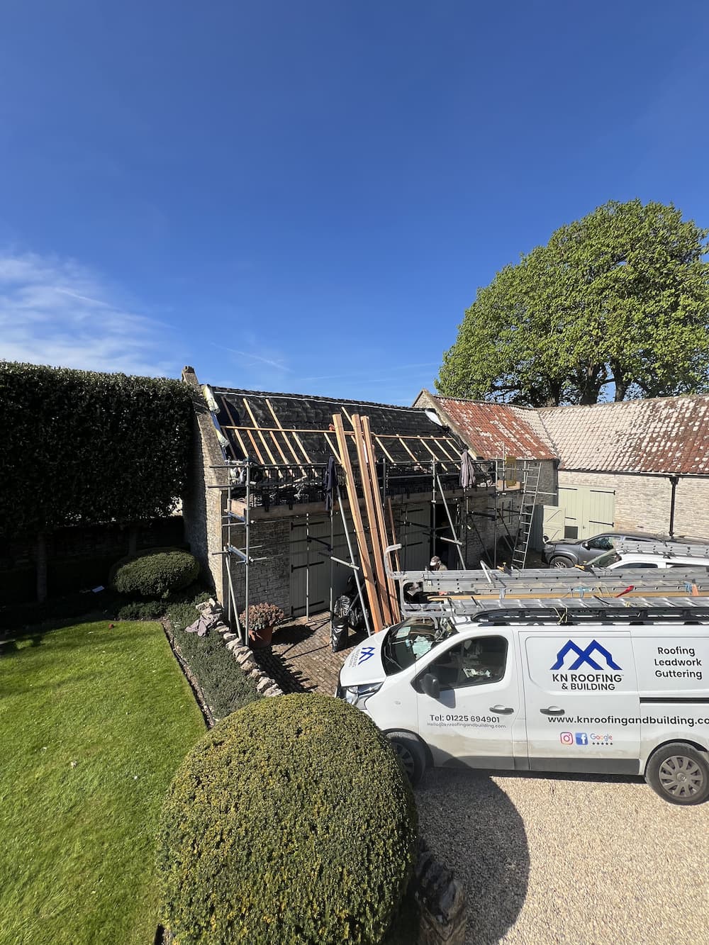 White Van with ladders on the roof parked in a gravel driveway next to a grassy garden and an old building with it's roof under construction