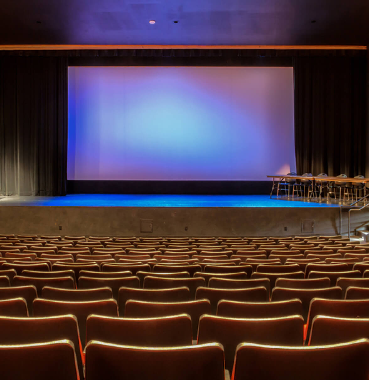 A spacious auditorium with rows of empty seats and a large blank screen illuminated in blue light.