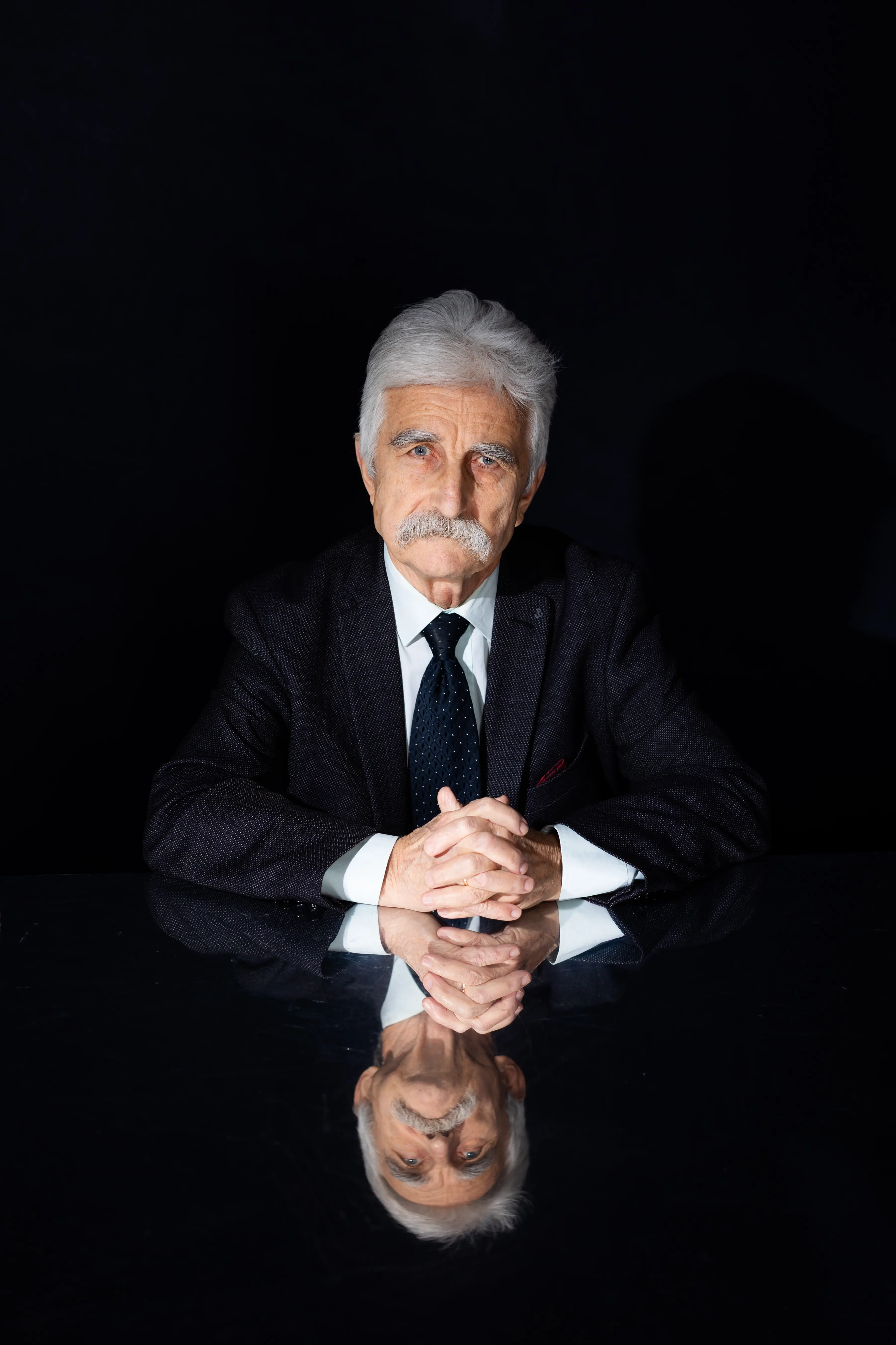 Starszy mężczyzna z siwymi włosami i wąsami siedzi przy czarnym stole ze splecionymi dłońmi, ubrany w ciemny garnitur i krawat. Jego odbicie jest widoczne na błyszczącej powierzchni stołu na czarnym tle.