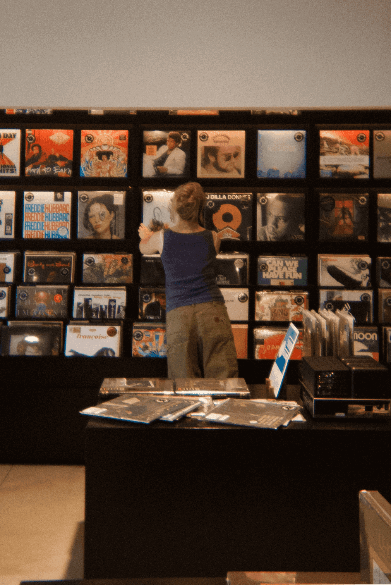 the wall of a record store with records lined up along the wall and a woman reaching to grab an album.