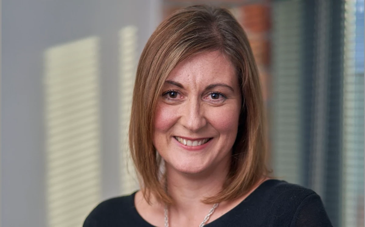 A person with shoulder-length light brown hair stands indoors against a softly lit background, wearing a dark top and a silver necklace.