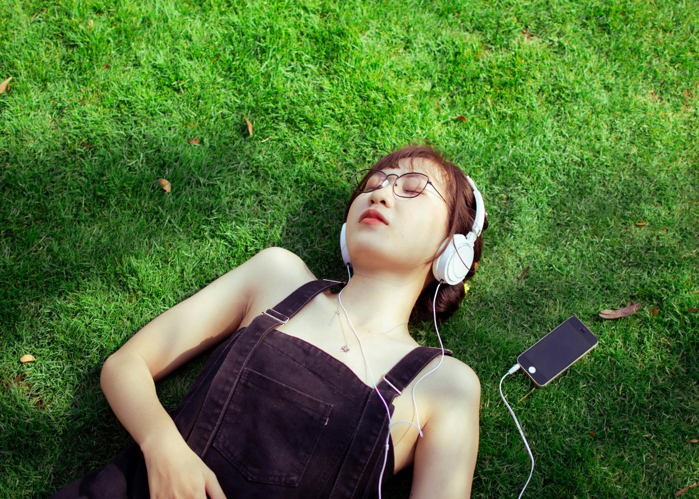 Woman with short hair and glasses laying down on grass listening to podcasts for sleep with her eyes closed