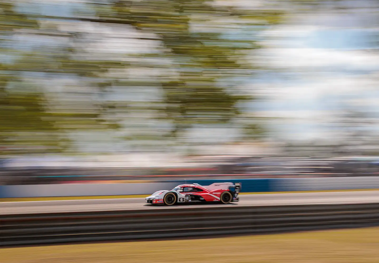 A red Porsche racing car speeds down a track, against a blue sky and trees, with a motion blurred background.