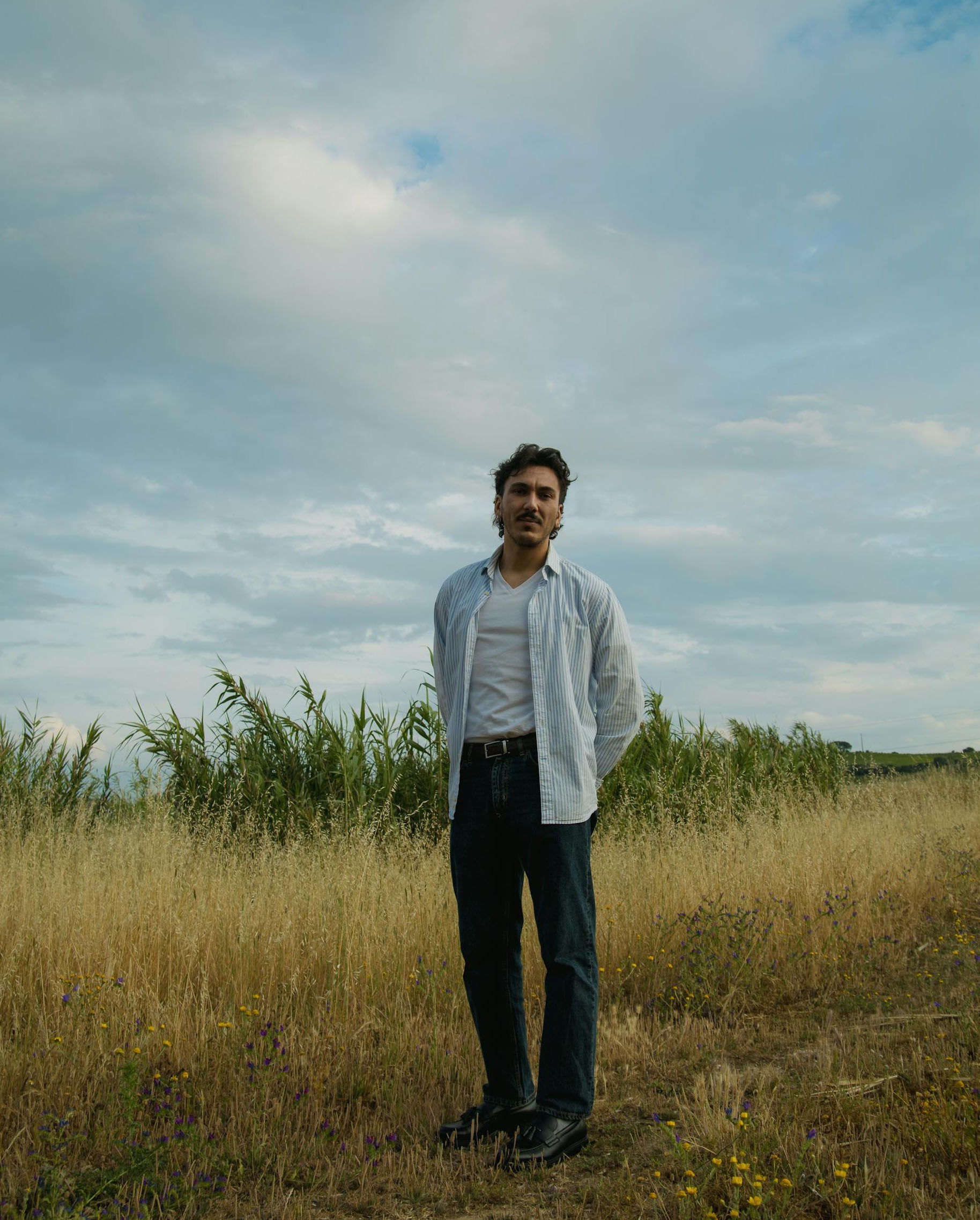 A man in an open shirt stands in a dry, grassy field