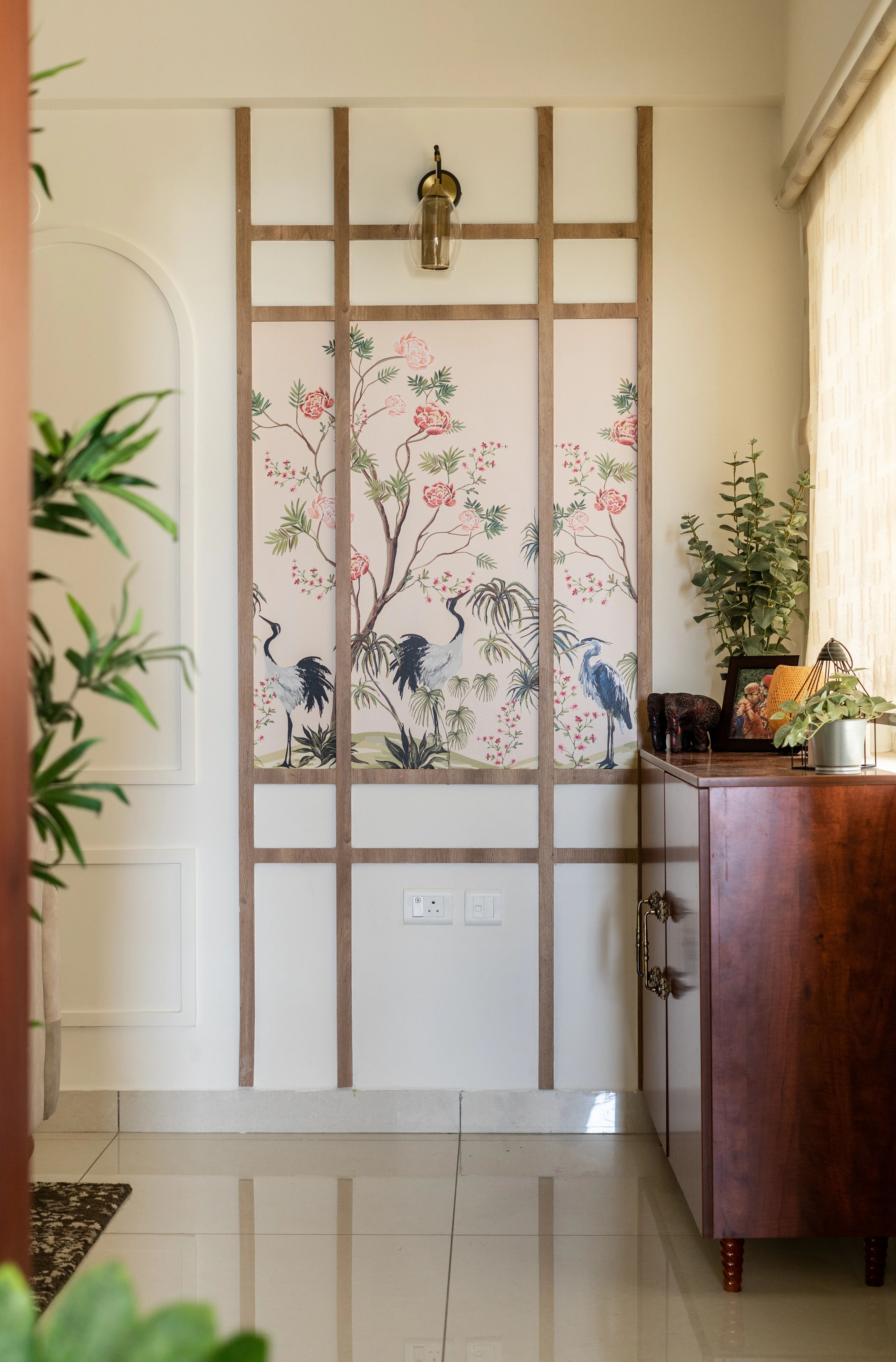 A foyer entryway featuring a traditional-style wallpaper with floral and bird motifs, framed by wooden beading. A wooden console table sits underneath, with potted plants on either side.