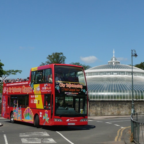 A red double-decker bus for city sightseeing drives past a large, dome-shaped glass conservatory on a sunny day.