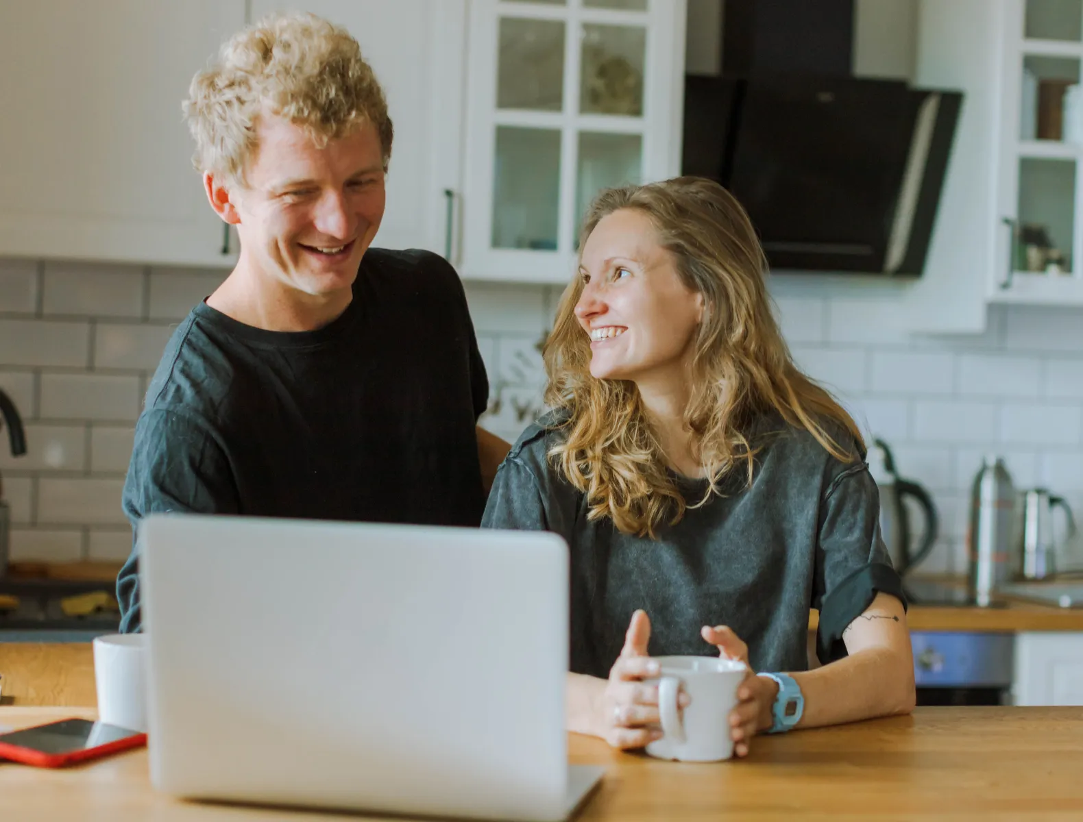 A couple in their kitchen smiling over morning coffee as they look at their computer.