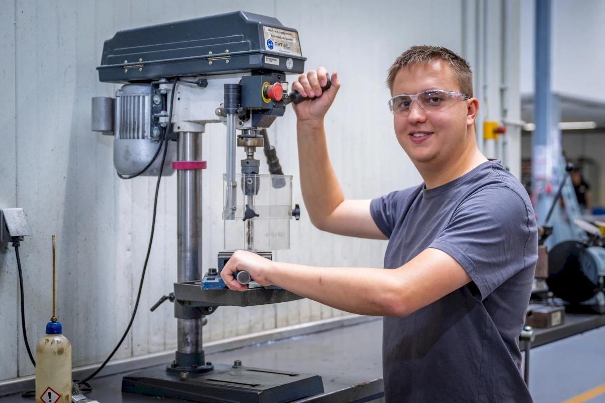 Mann benutzt eine Standbohrmaschine mit einer Schutzbrille in einer Werkstatt. Er lächelt, während er die Maschine bedient.
